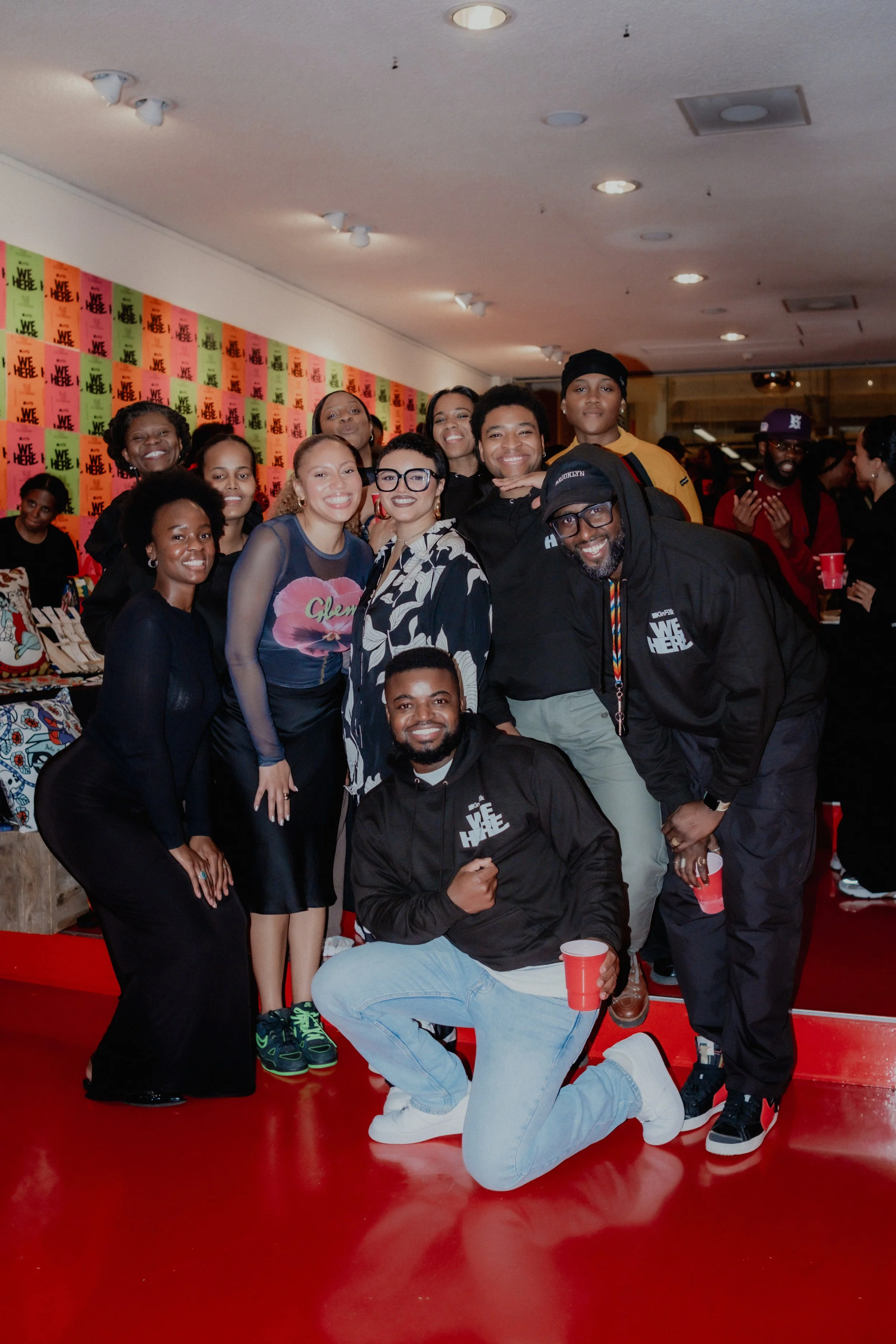 Group of diverse people posing together at an indoor event, some holding red cups, with a colorful backdrop of posters and other attendees in the background.