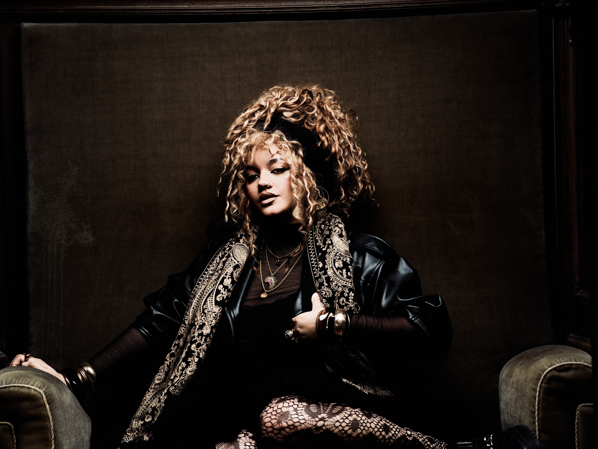 A woman with curly hair, wearing a black leather jacket and fishnet stockings, sitting on a green velvet sofa with a dark background.