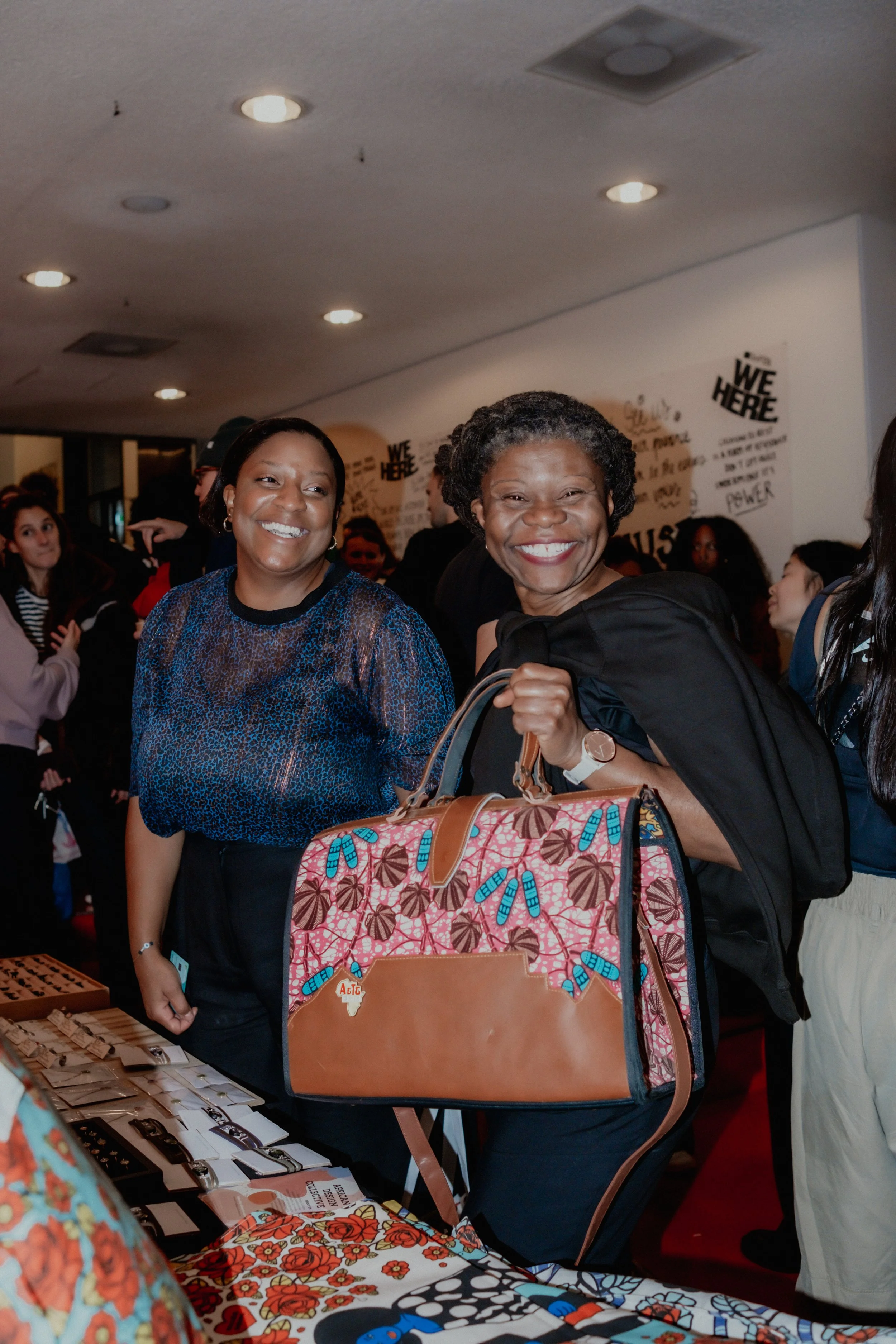 Two women smiling at a social event, surrounded by other people, with display tables and artwork on the wall.