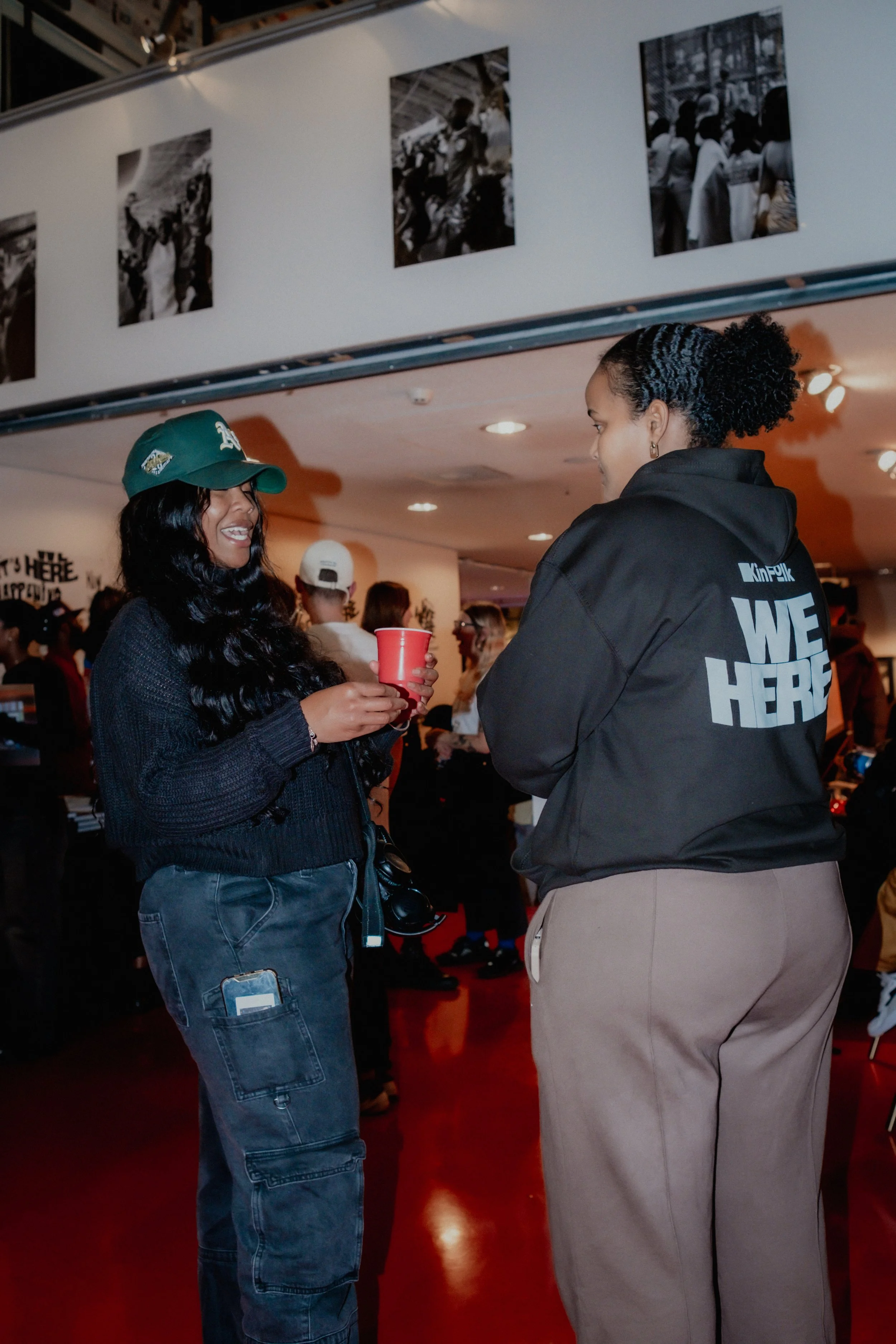 Two women conversing at an indoor event, one is Asian with long black hair and glasses, wearing a dark jacket and a green cap, holding a red cup. The other woman is African American with curly black hair tied back, wearing a black hoodie with white t