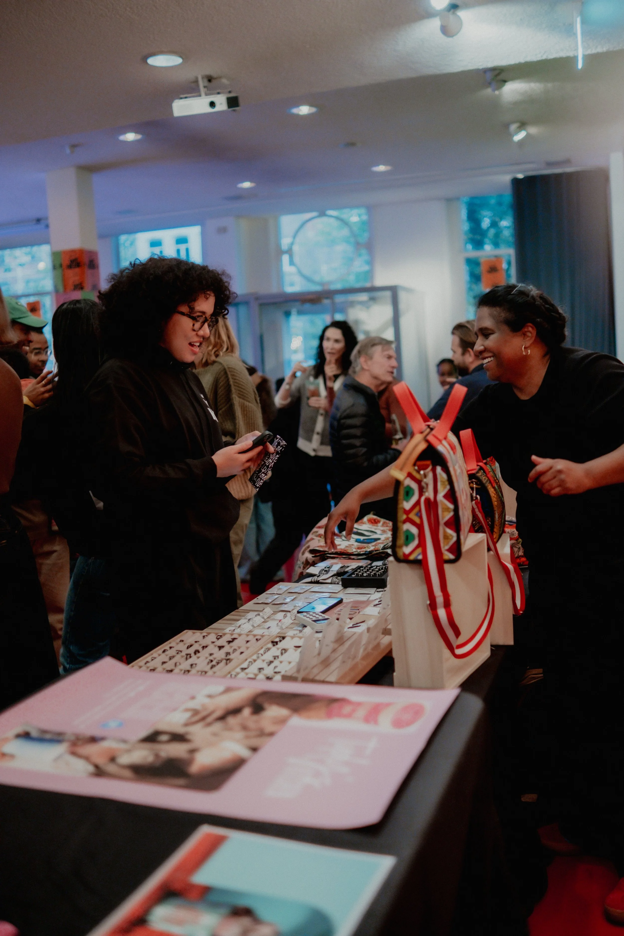 People browsing and shopping at a market stall inside a store or event space, with tables displaying various items, including bags with colorful patterns and magazine or catalog pages on the table in the foreground.