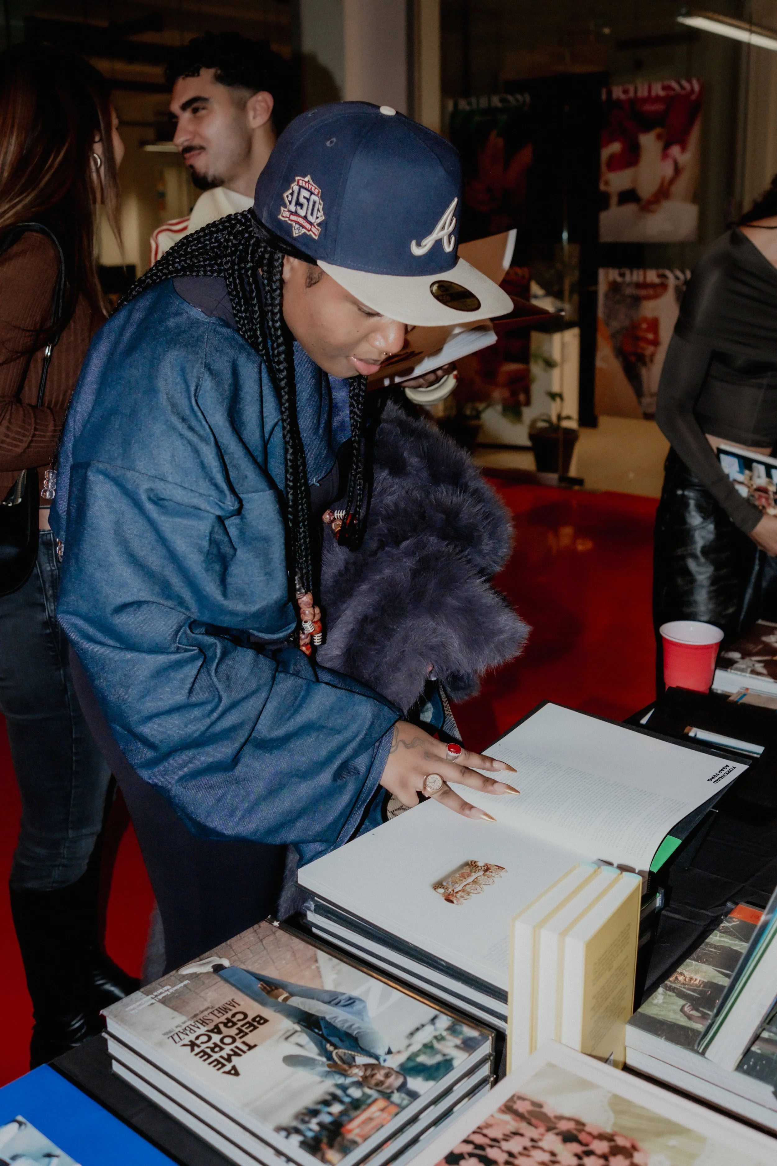 A person with braided hair wearing a baseball cap and denim jacket looking at a book on display table at a book event.