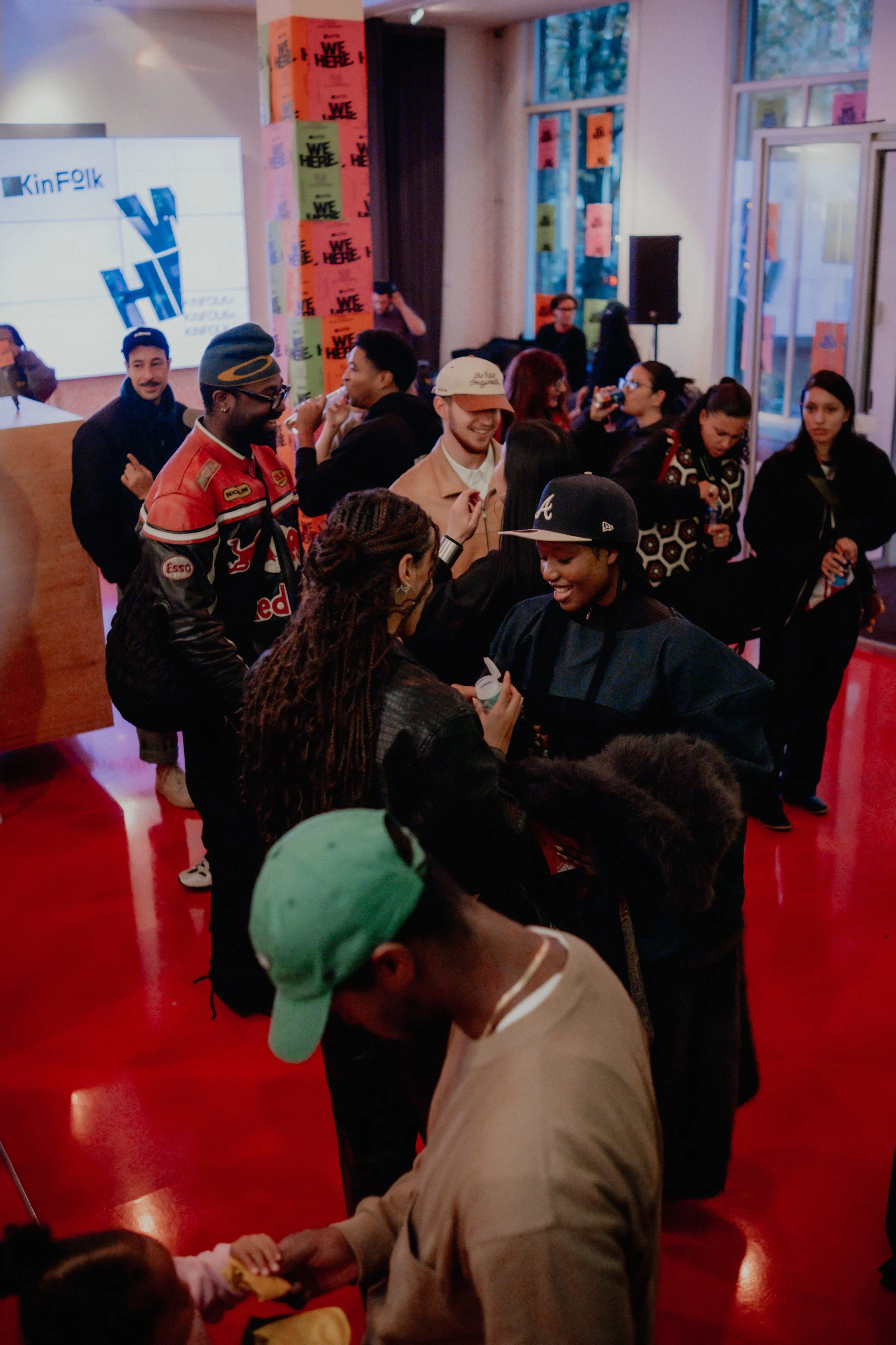 A group of people gather indoors during an event, some are smiling, talking, and taking photos. The setting appears to be a vibrant, lively space with colorful posters on the walls and a large screen displaying a logo that reads 'KinFolk'.
