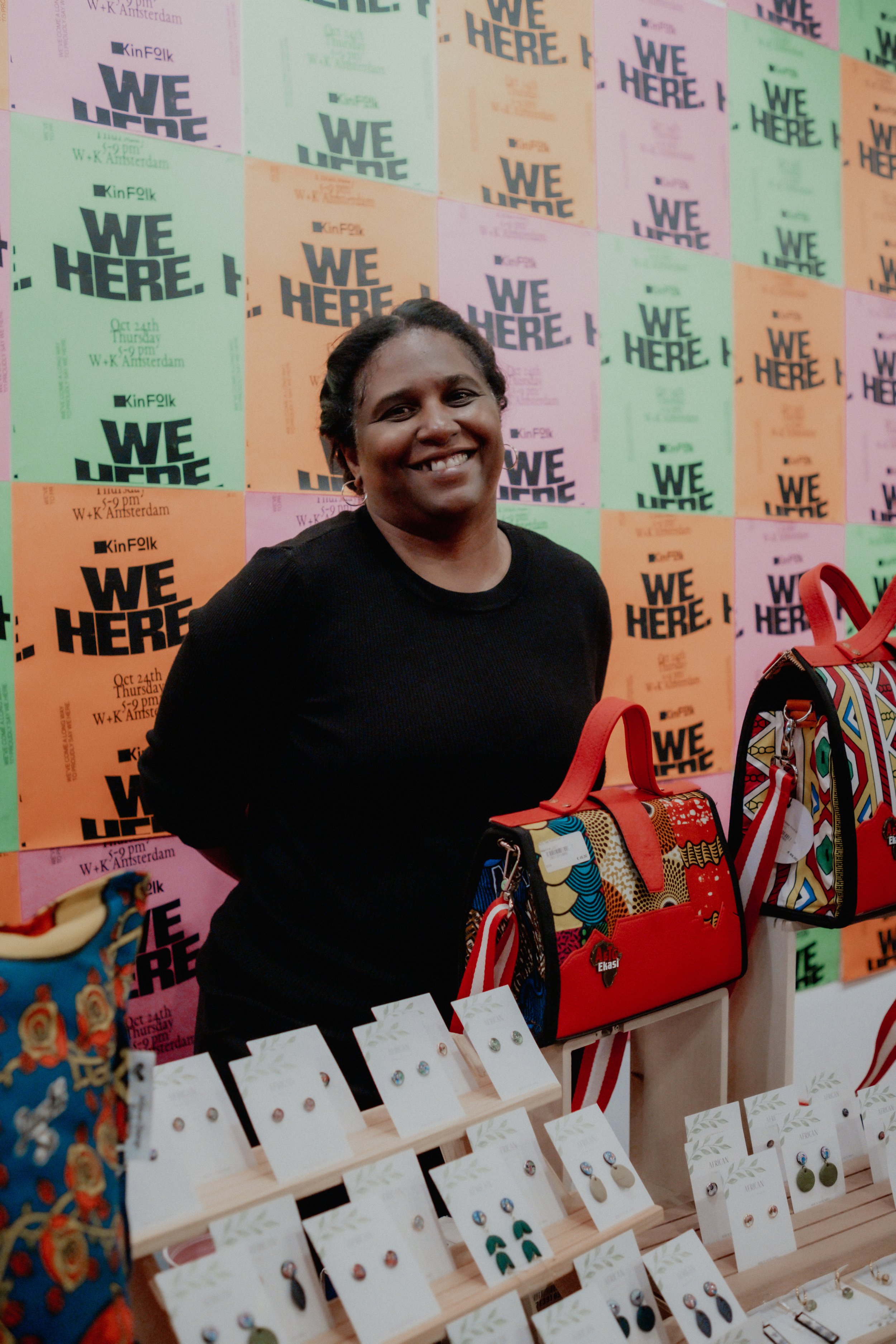 A woman smiling in front of a colorful background with promotional posters that read 'WE ARE HERE'. In front of her is a display of earrings on white cards, and there are colorful handbags on display.