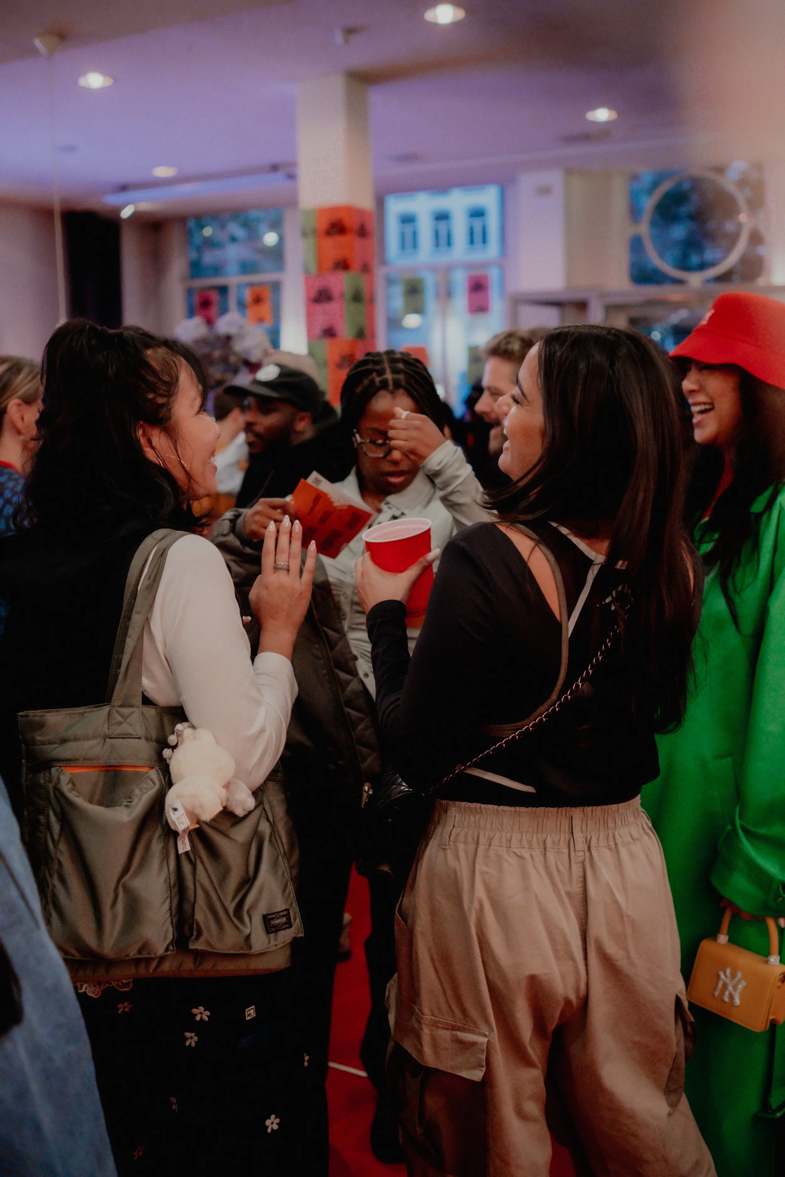 Group of diverse people in a crowded indoor space, engaging in conversation and laughing, with colorful posters on the walls.
