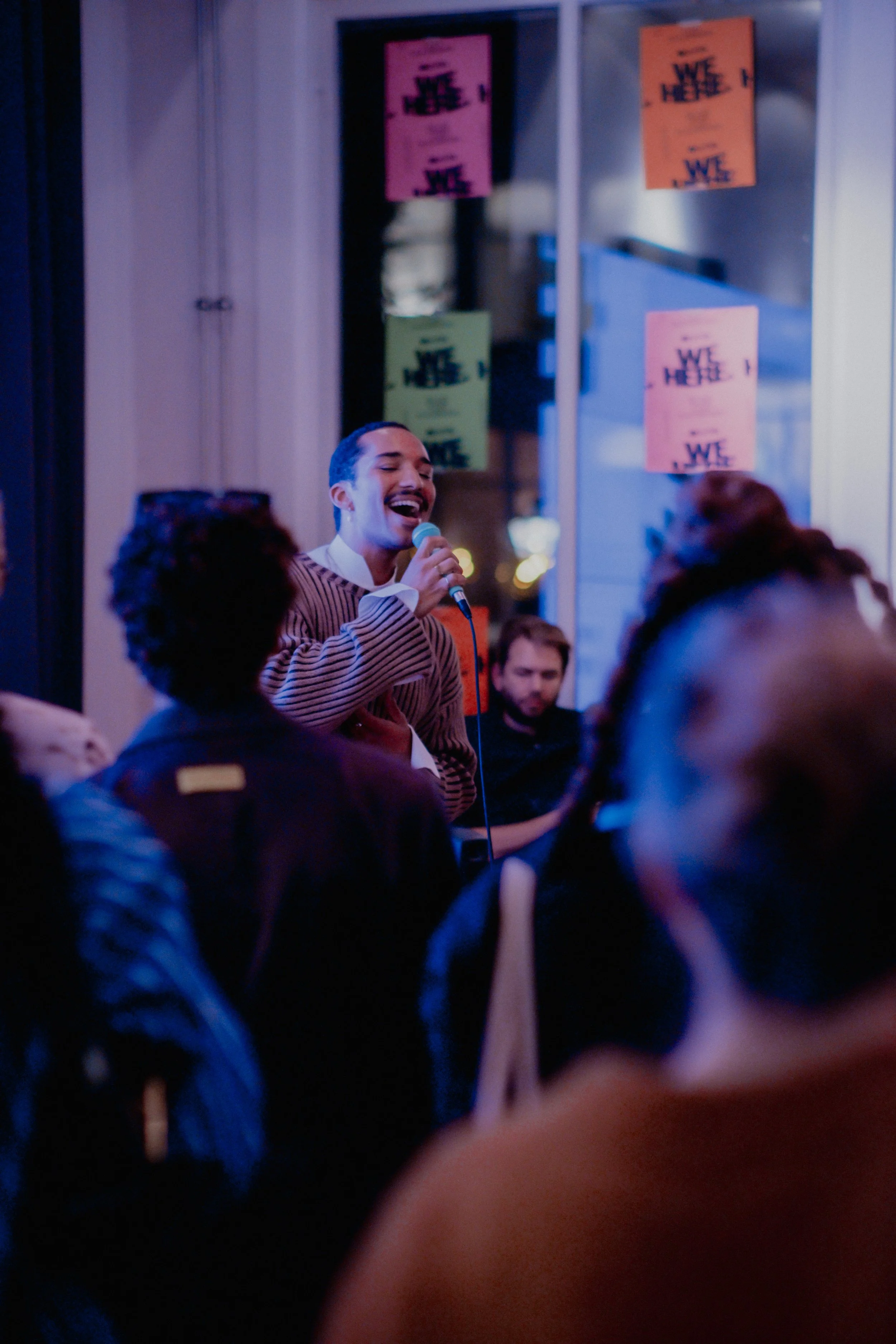 A man with long dark hair singing into a microphone at an indoor event, with people and colorful posters on the window in the background.