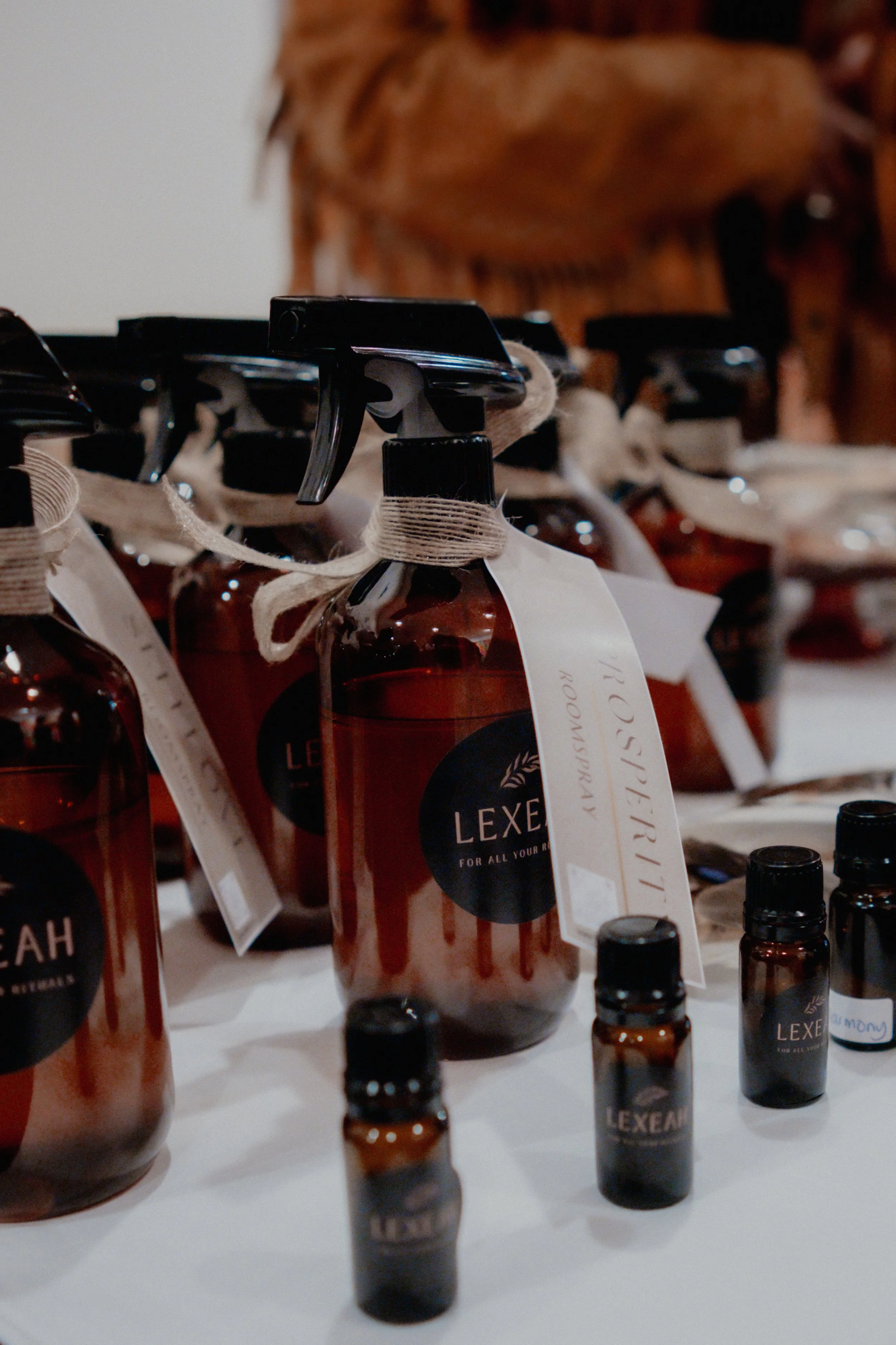 Close-up of several small brown glass bottles with black caps and labels, some with white tags tied around their necks, arranged on a white surface with a blurred background.