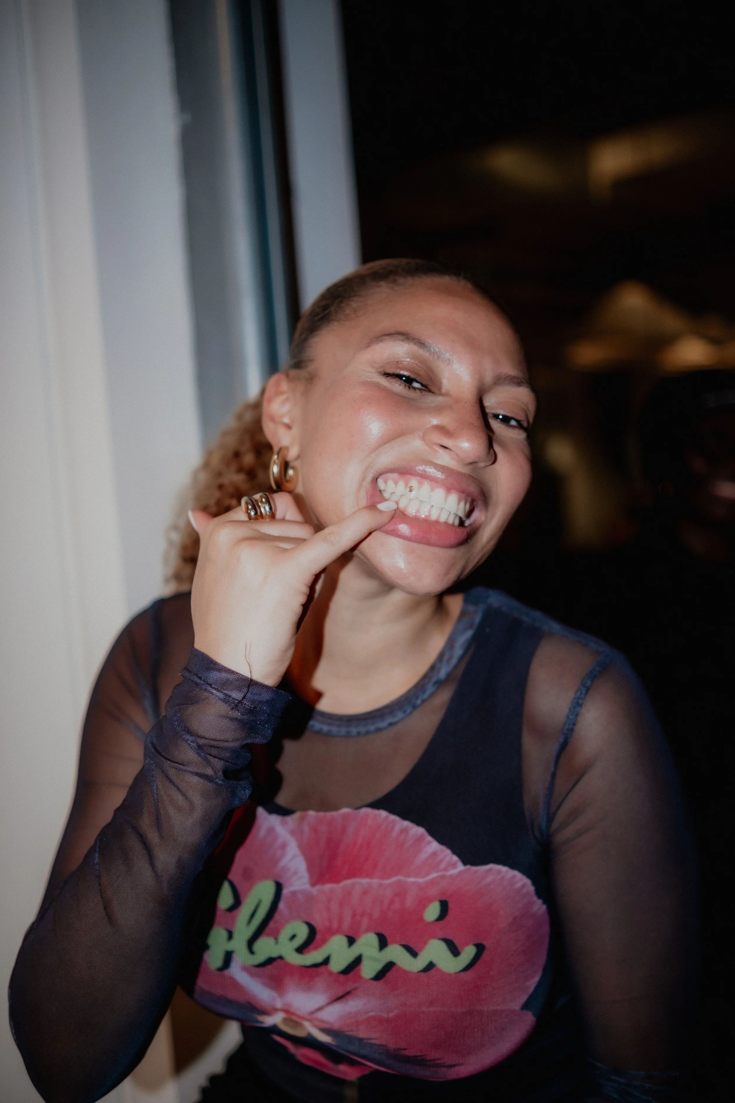 A woman smiling widely, showing her teeth, with one hand touching her face. She is wearing hoop earrings, a sheer black long sleeve top, and a black T-shirt with pink text.