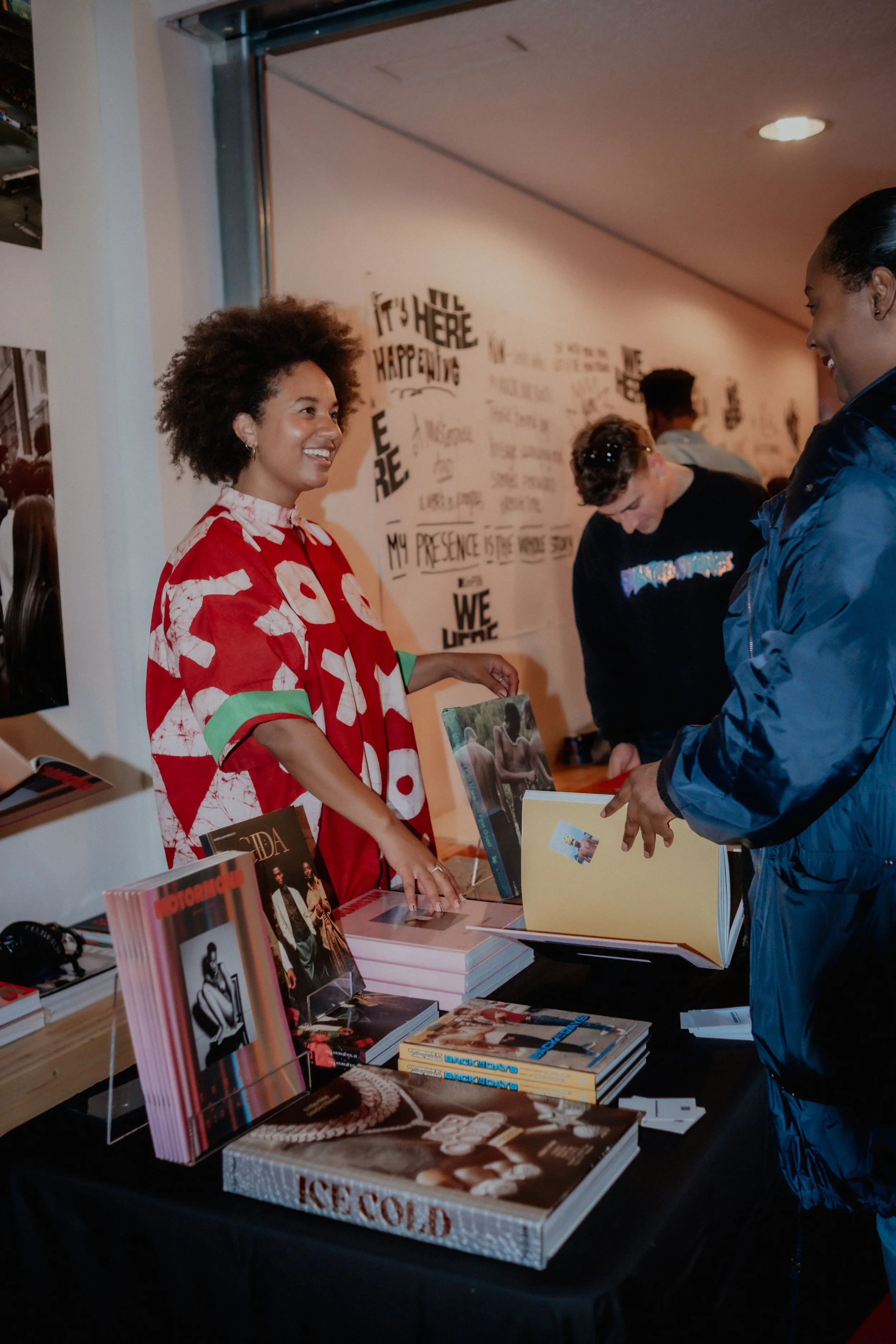 A woman in a red and white patterned shirt smiling and talking to a man in a blue jacket at a book signing event. There are several books displayed on the table, including one titled 'Ice Cold,' and colorful posters on the wall behind them.