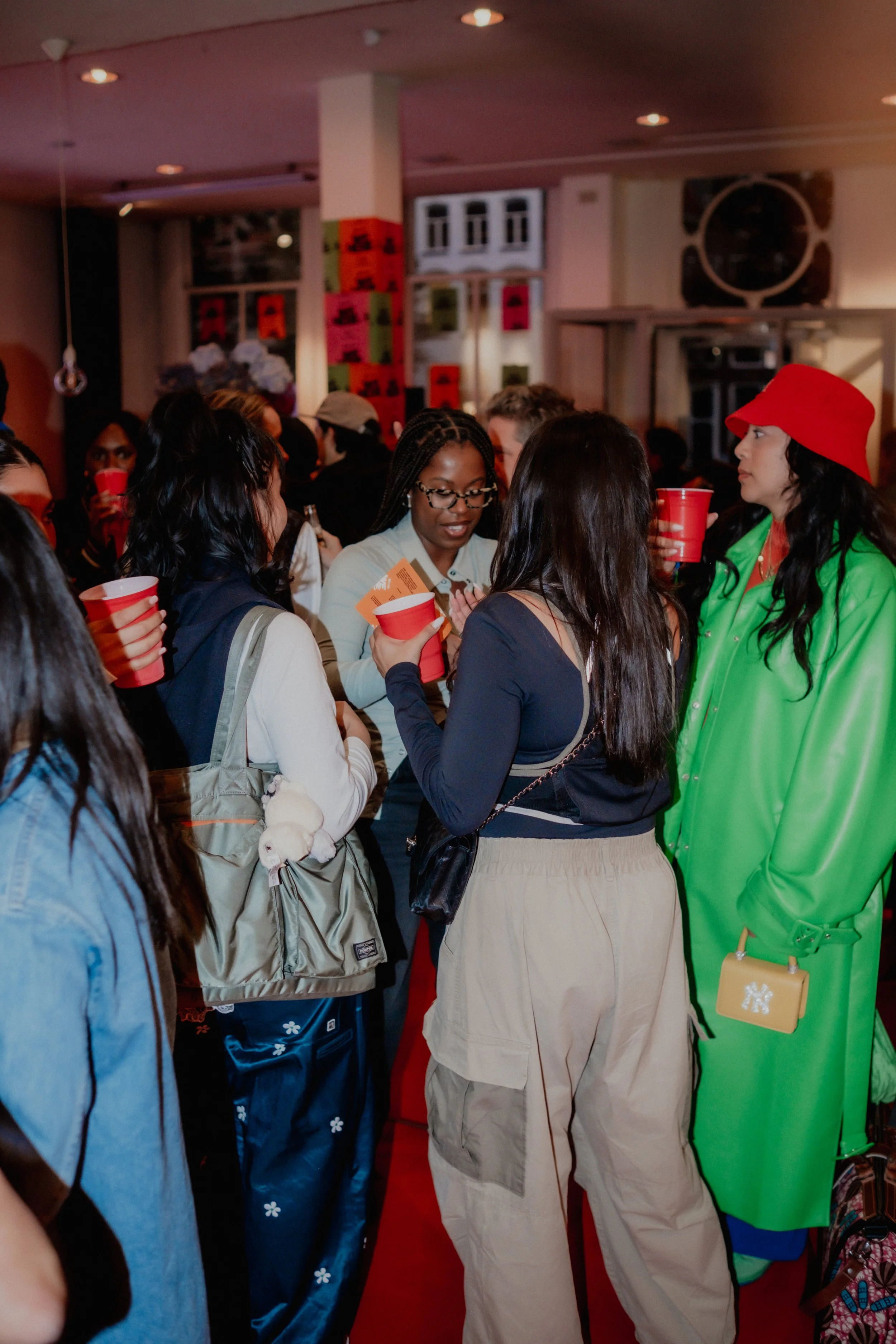 Group of people at an indoor social event, some holding red cups, engaged in conversations. A woman in a bright green outfit and red hat stands out. The background has colorful posters and a set of large speakers.