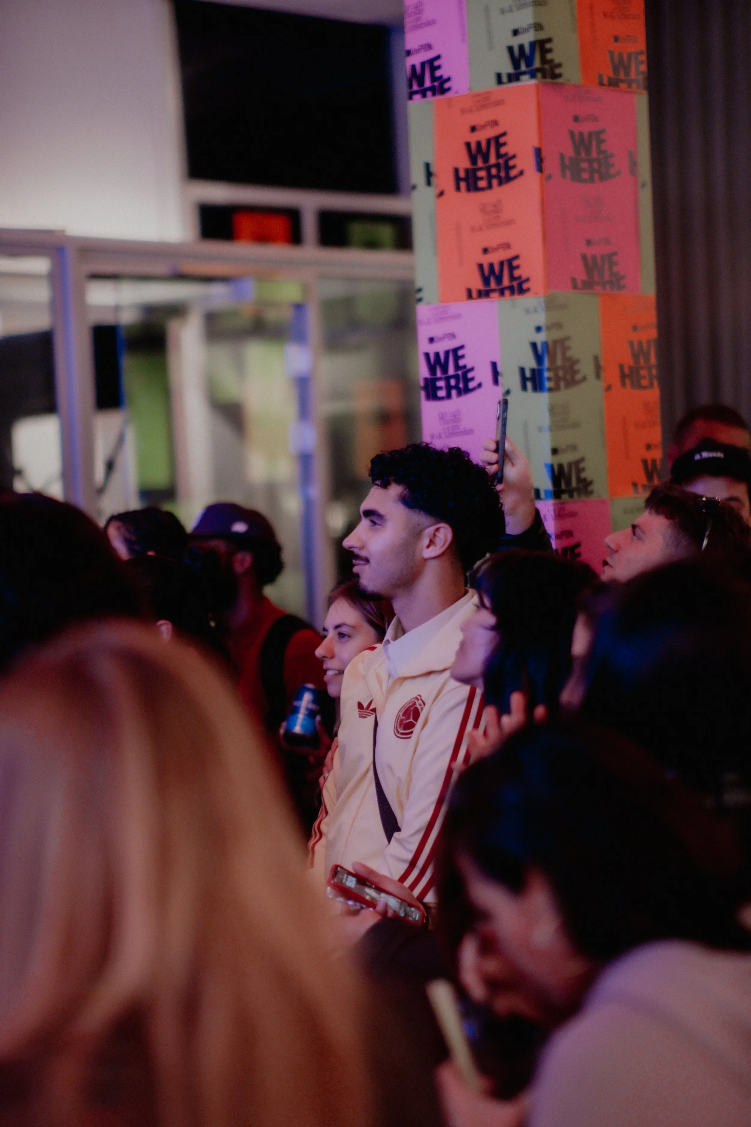 A crowd of people at an indoor event with colorful posters in the background that read 'WE HERE'. A young man with curly hair and a light-colored jacket stands among the crowd, holding a smartphone.