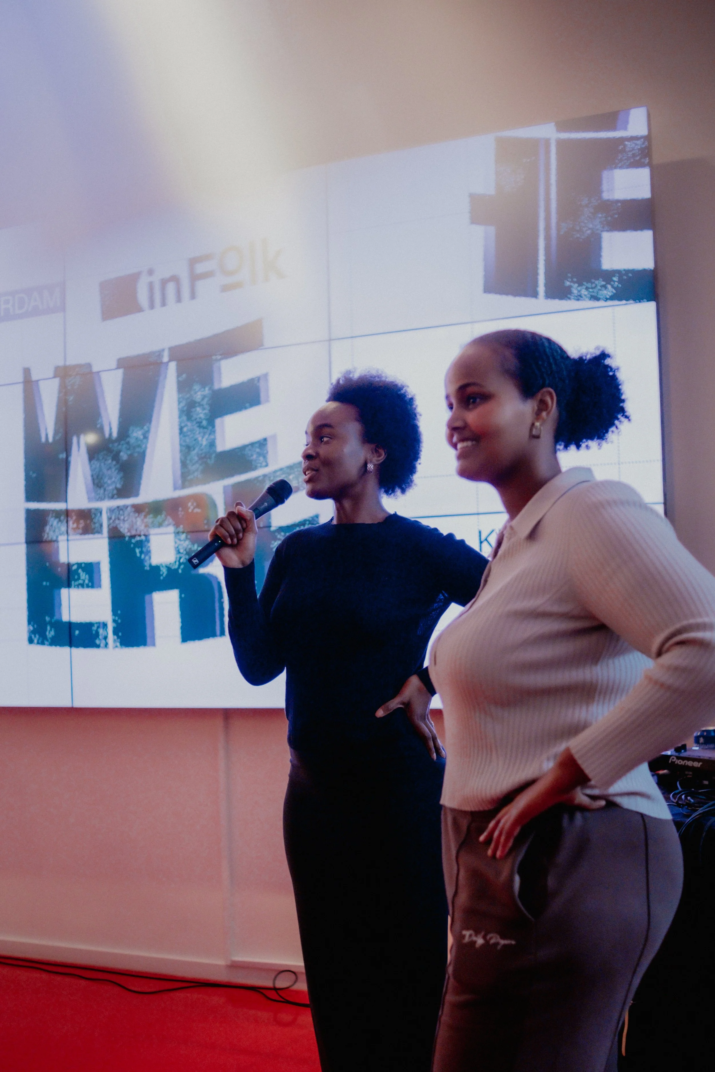 Two women standing together in front of a large screen, one holding a microphone, at an indoor event or presentation.