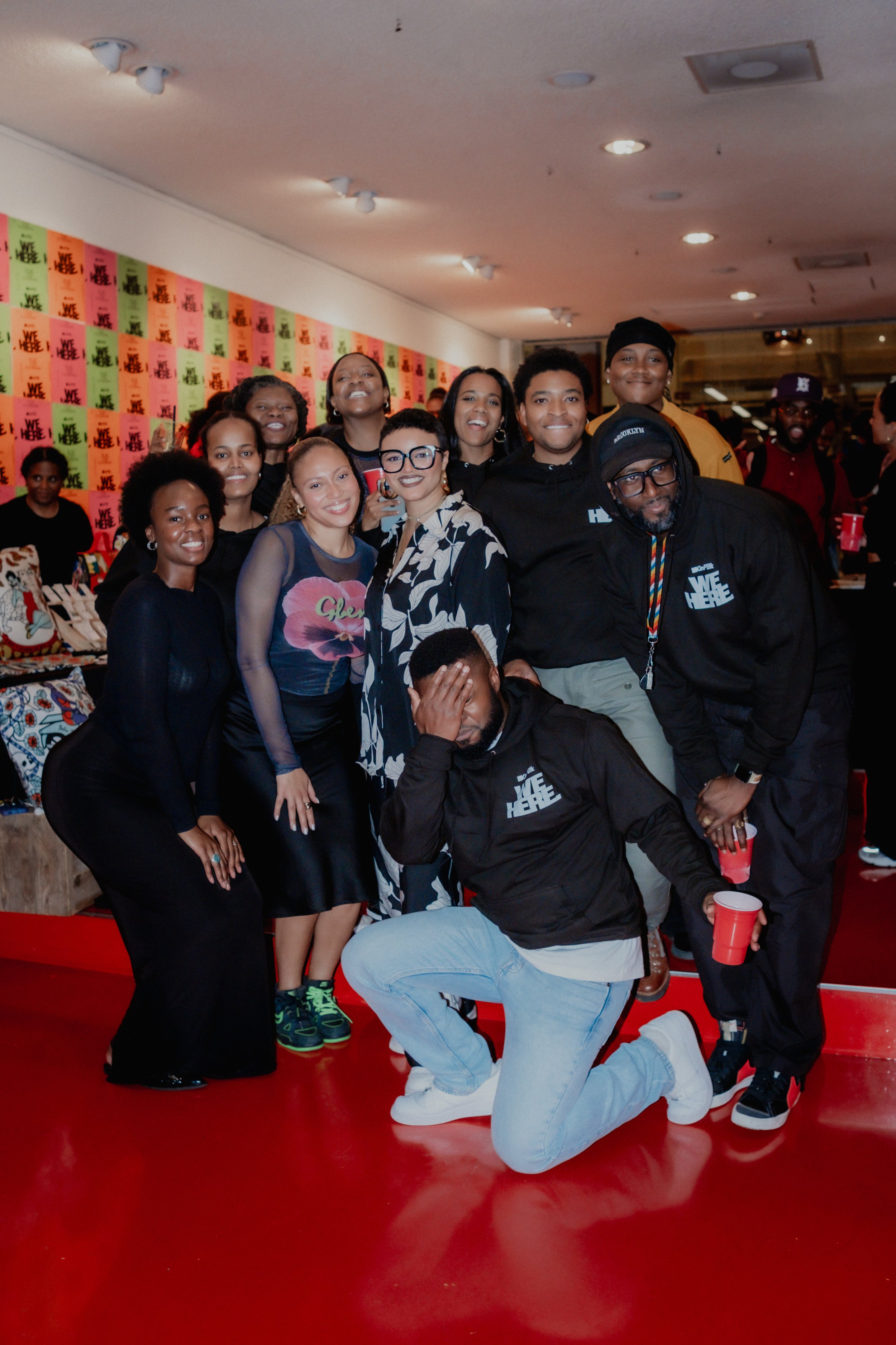 A group of people smiling and posing for a photo at an indoor event with a wall covered in colorful posters that say 'WE HERE.' Some are holding red cups, and one person is kneeling in front with hand covering their face.