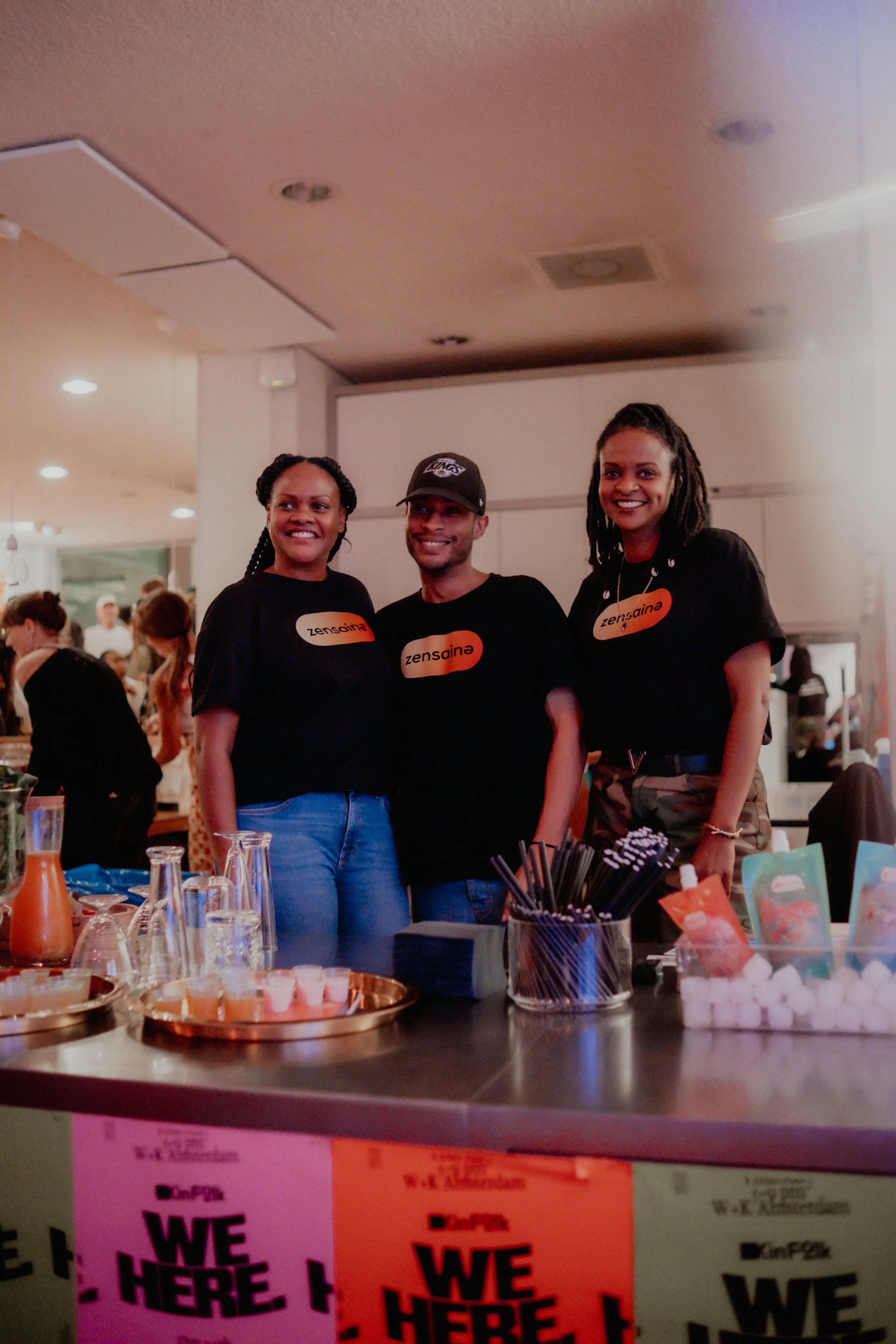 Three women smiling and standing behind a table at an indoor event, wearing black t-shirts with the word 'zensaina' on them.