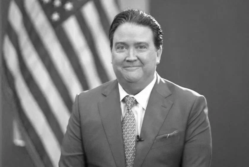 A man in a suit smiling in front of an American flag.