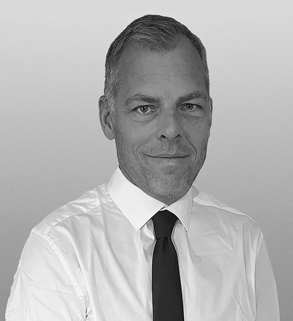 Black and white headshot of a man with short hair, wearing a white shirt and black tie, smiling slightly, against a plain background.