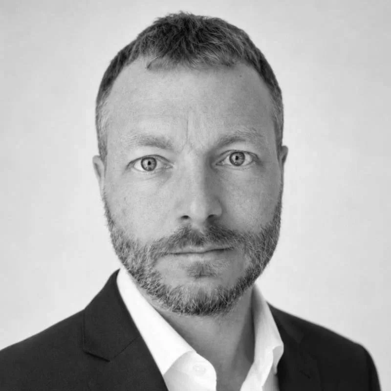 Black and white headshot of a man with short hair and a beard, wearing a suit jacket and white shirt, looking directly at the camera.