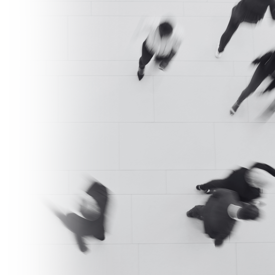 Black and white photo of six people walking in a large open indoor space, taken from above. The people are blurred, indicating movement.