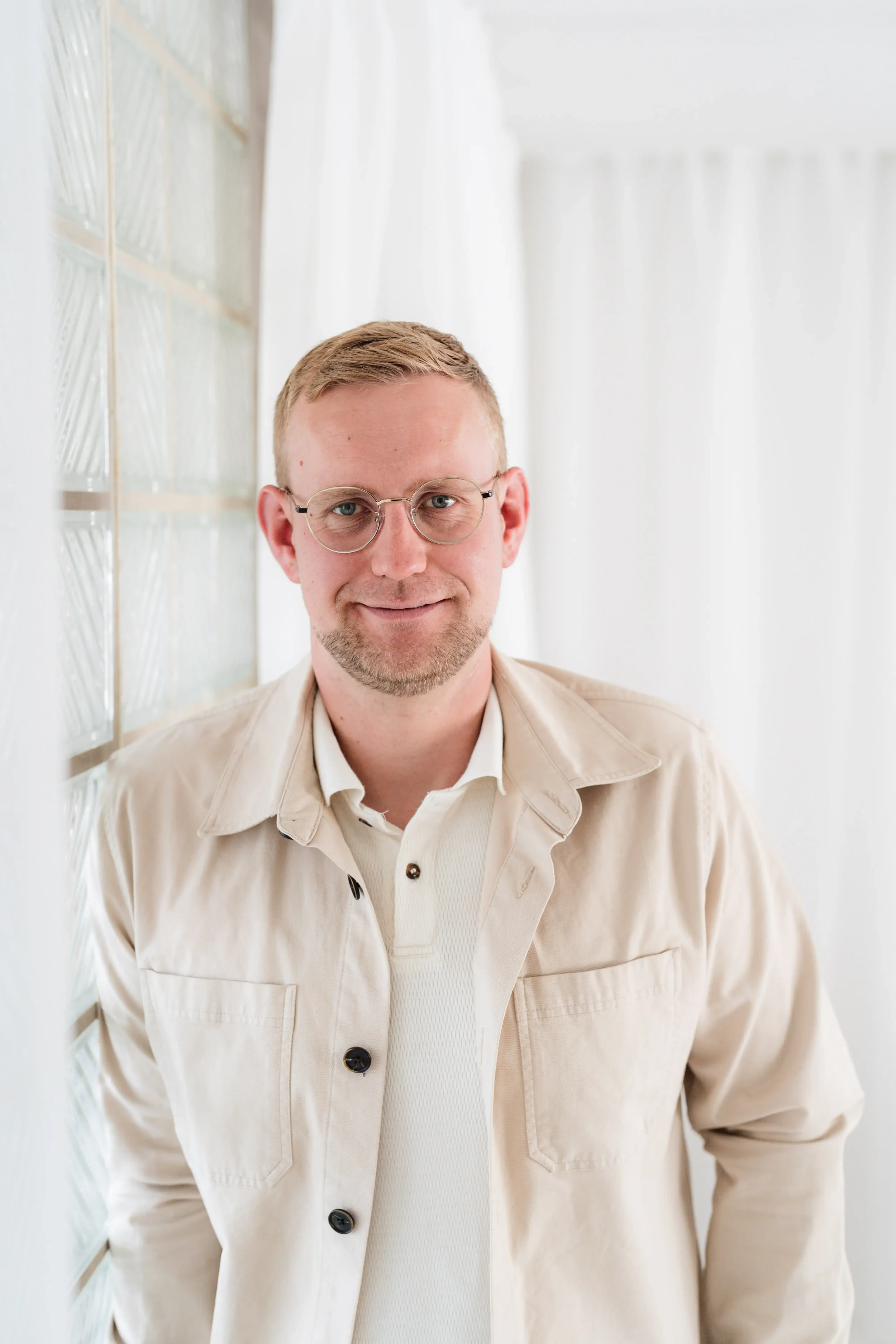 Ein Mann trägt eine beige Jacke und eine weiße Hemd, trägt runde Brille und lächelt vor weißem Hintergrund.