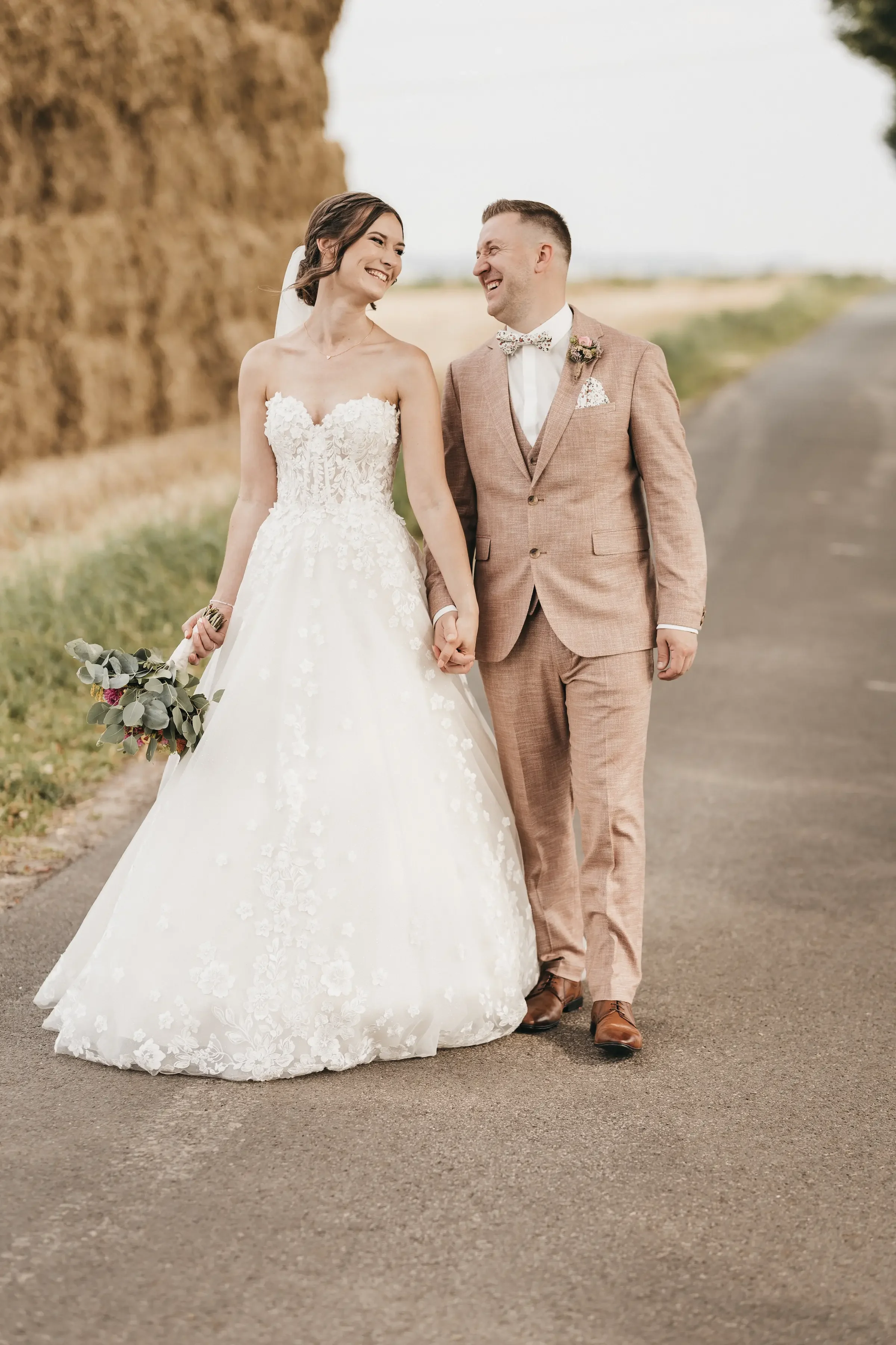 Ein Brautpaar hält Händchen und lächelt sich auf einer Straße mit Naturkulisse im Hintergrund. Das Brautkleid ist lang und weiß mit floraler Spitze, der Bräutigam trägt einen braunen Anzug mit Fliege.
