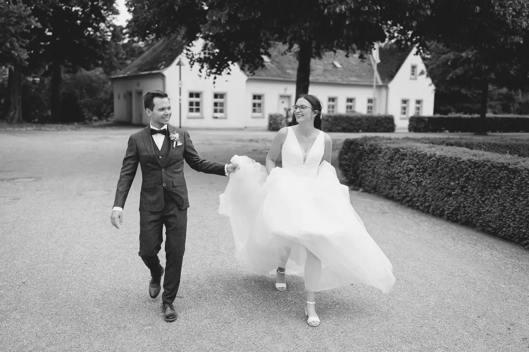 Ein glückliches Paar in Hochzeitskleidung läuft draußen auf einem Weg, umgeben von Büschen und Bäumen, im Hintergrund ein Haus.