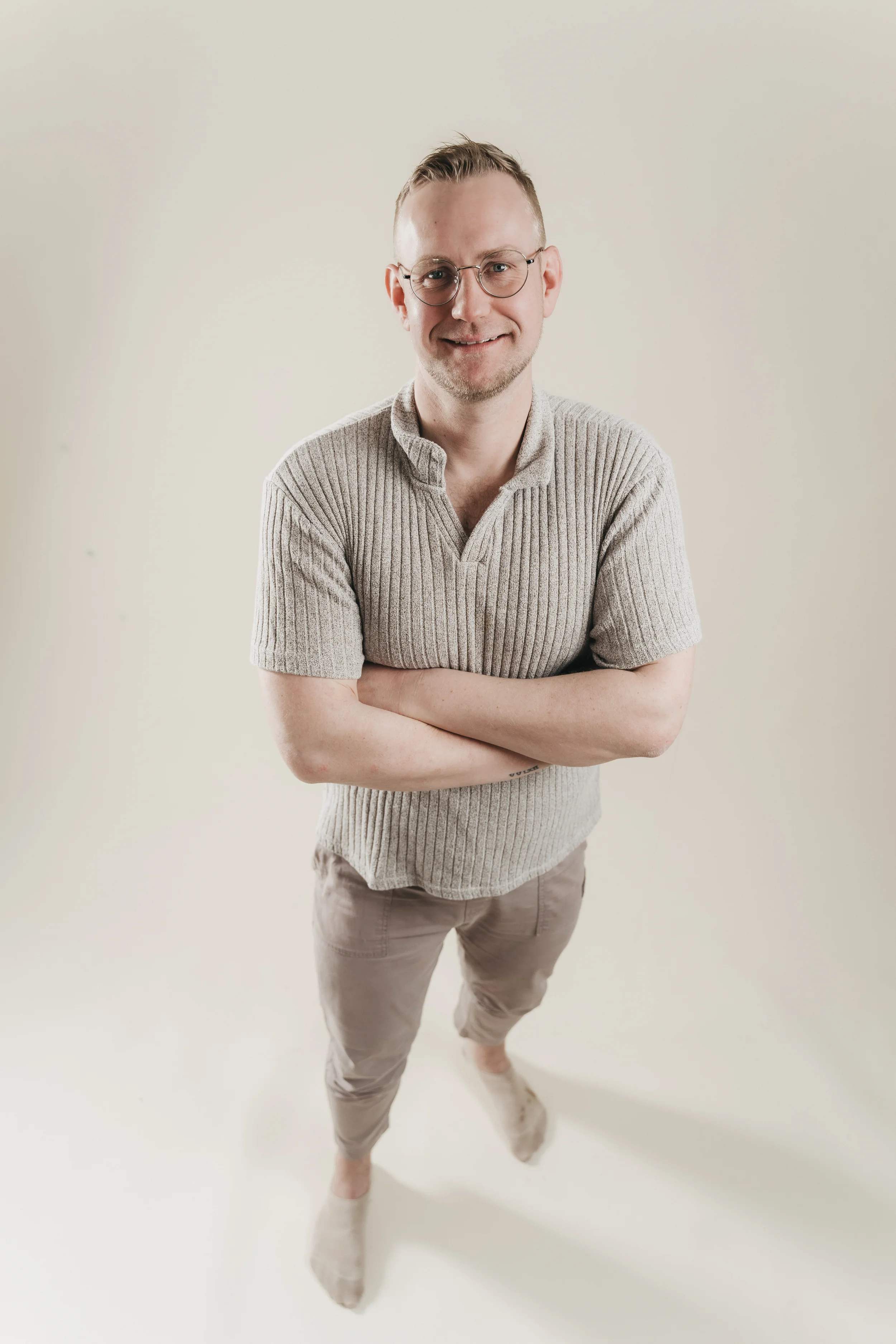 Junge Person mit Brille trägt beige Pullover und Hellbraune Hosen, steht vor neutralem Hintergrund, lächelt, Arme verschränkt.