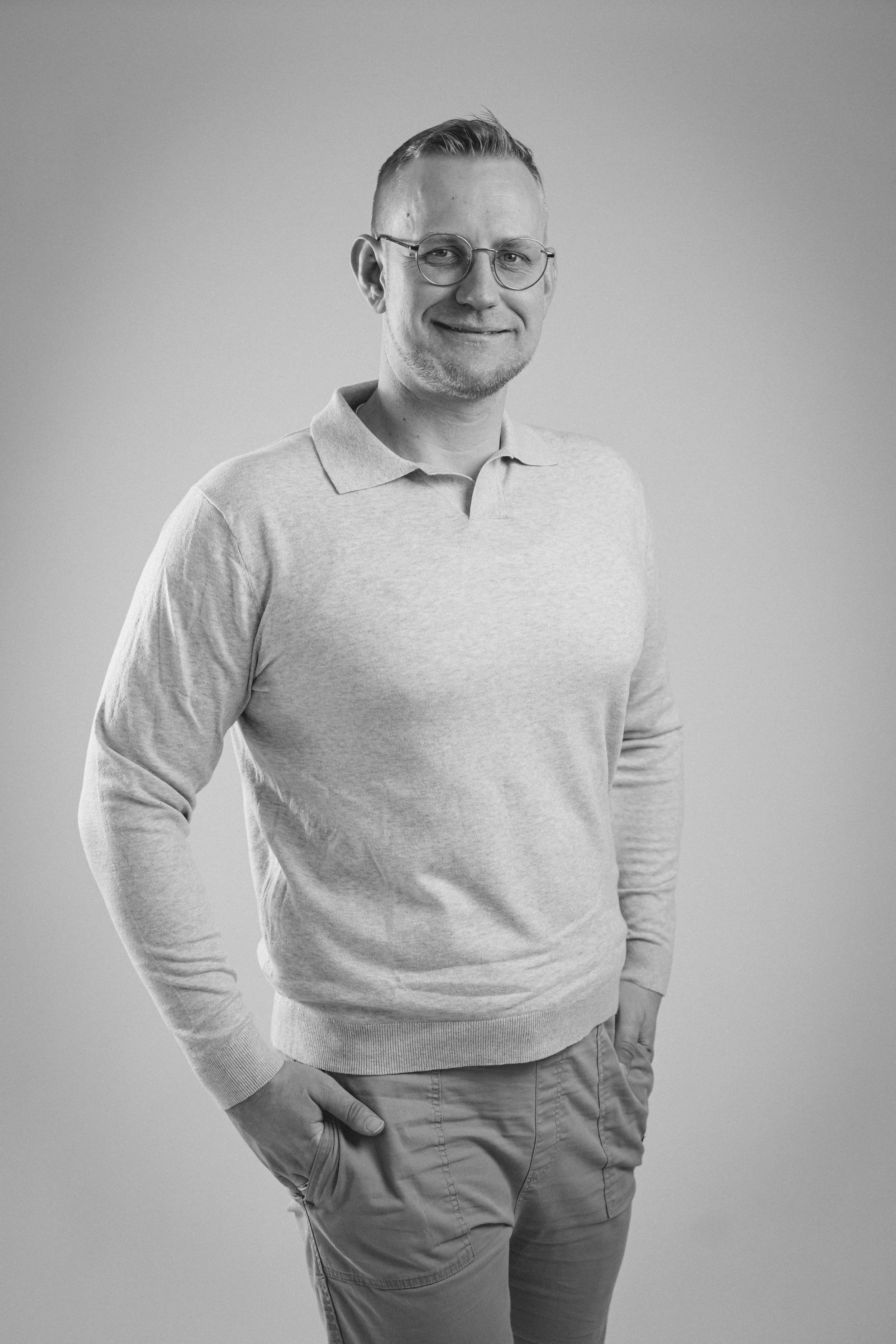 Schwarz-weiße Porträtaufnahme eines lächelnden Mannes, der eine Brille trägt, in einem hellen Oberteil und Kordhose, mit einer Hand in der Tasche, vor hellem Hintergrund.