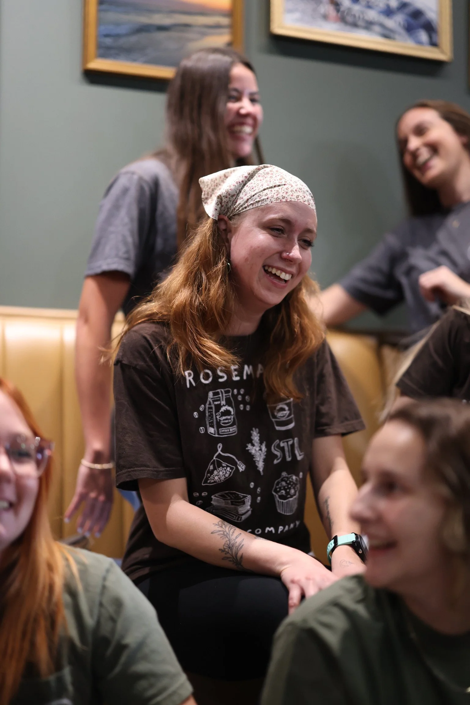 Group of young women laughing and smiling at a social gathering inside a room decorated with framed paintings on the wall.