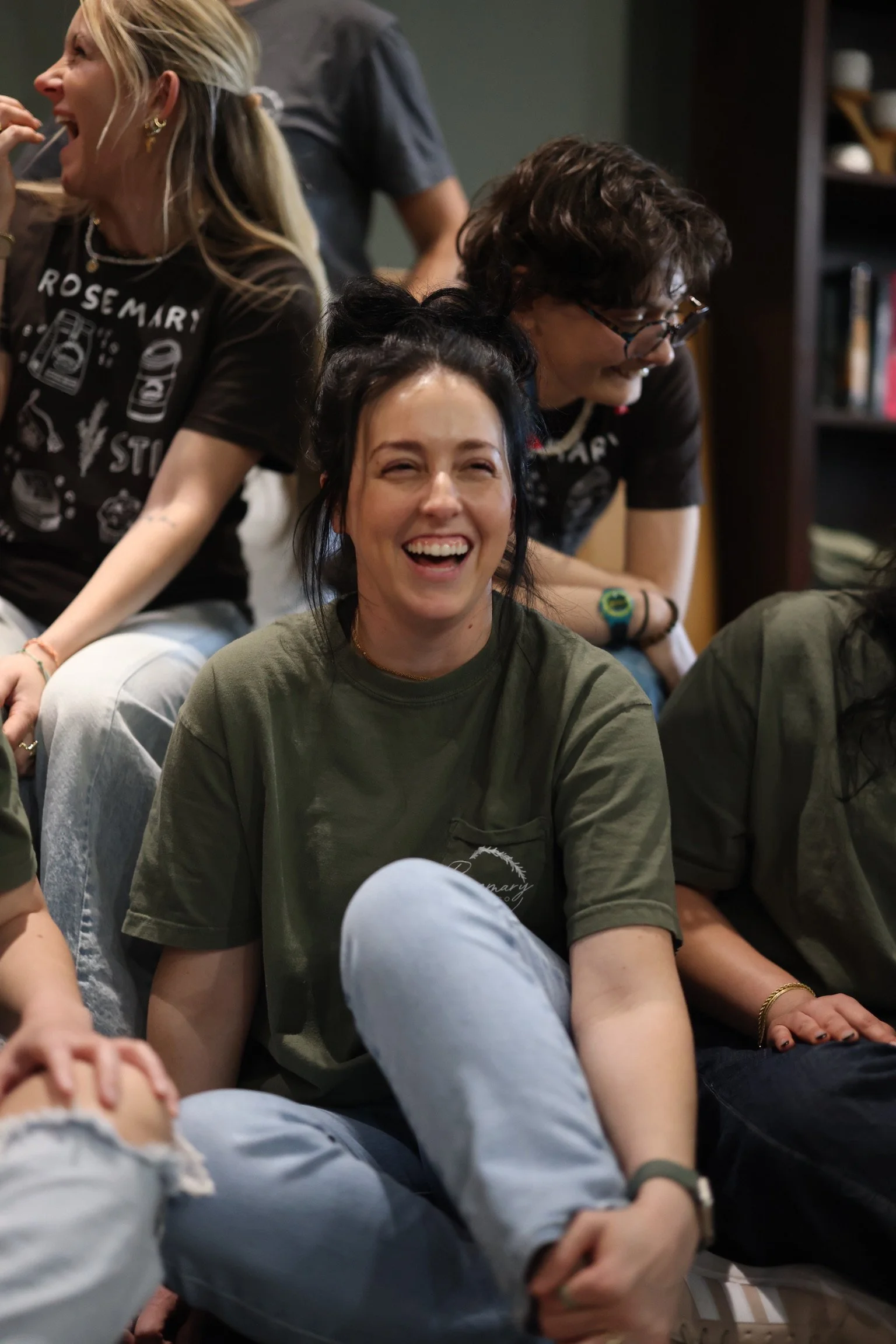 Group of young adults laughing and sitting closely together indoors, with bookshelves in the background.
