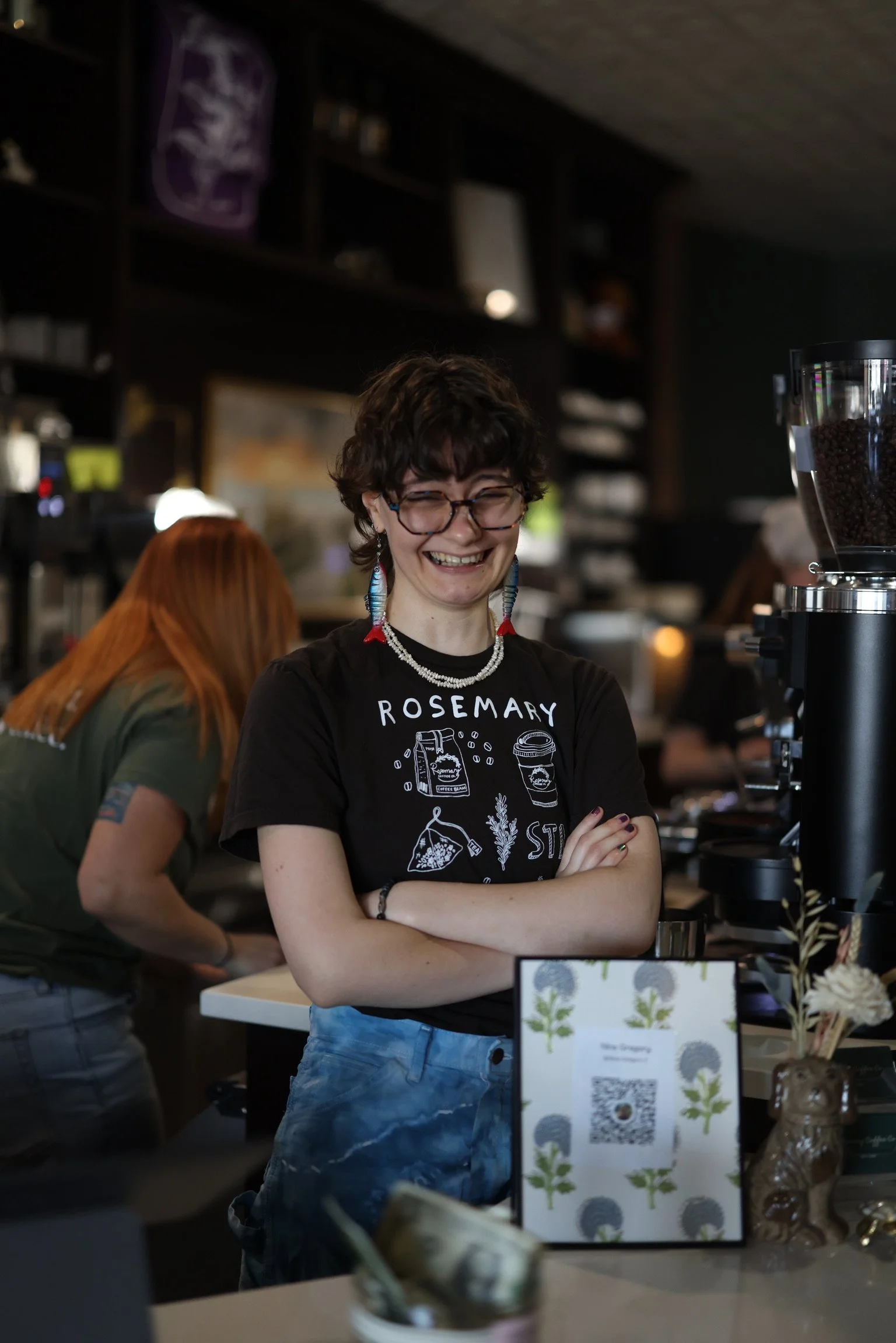 A young woman with short curly hair, glasses, and colorful earrings smiling at a cafe or shop. She is wearing a black t-shirt with the word 'ROSEMARY' and illustrations of herbs and coffee. She is standing with arms crossed near a display table with a botanical poster, coffee grinder, and other items.