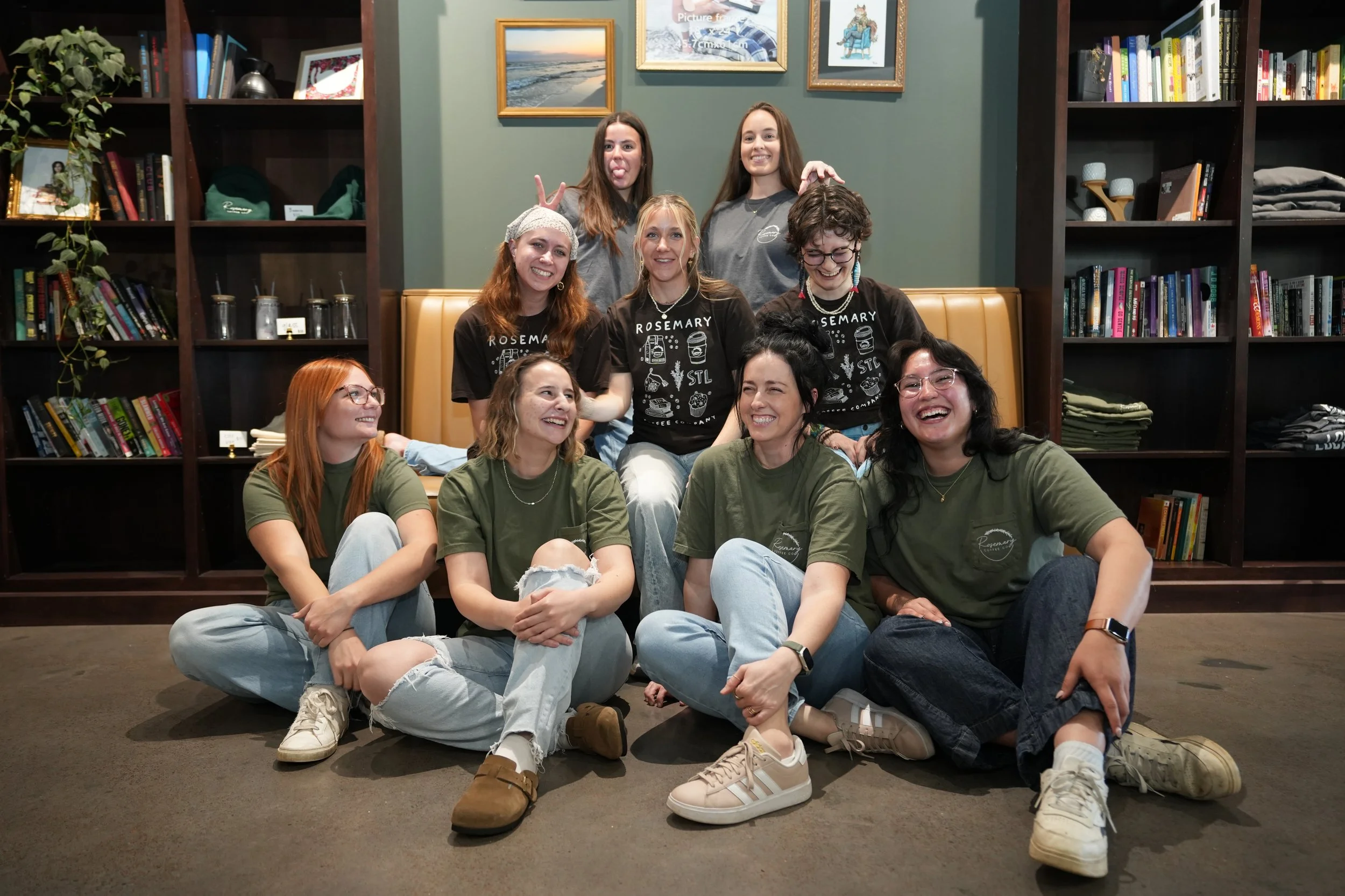 Group of nine young women sitting and standing in front of bookshelves, posing playfully with smiles and gestures in a cozy indoor setting.