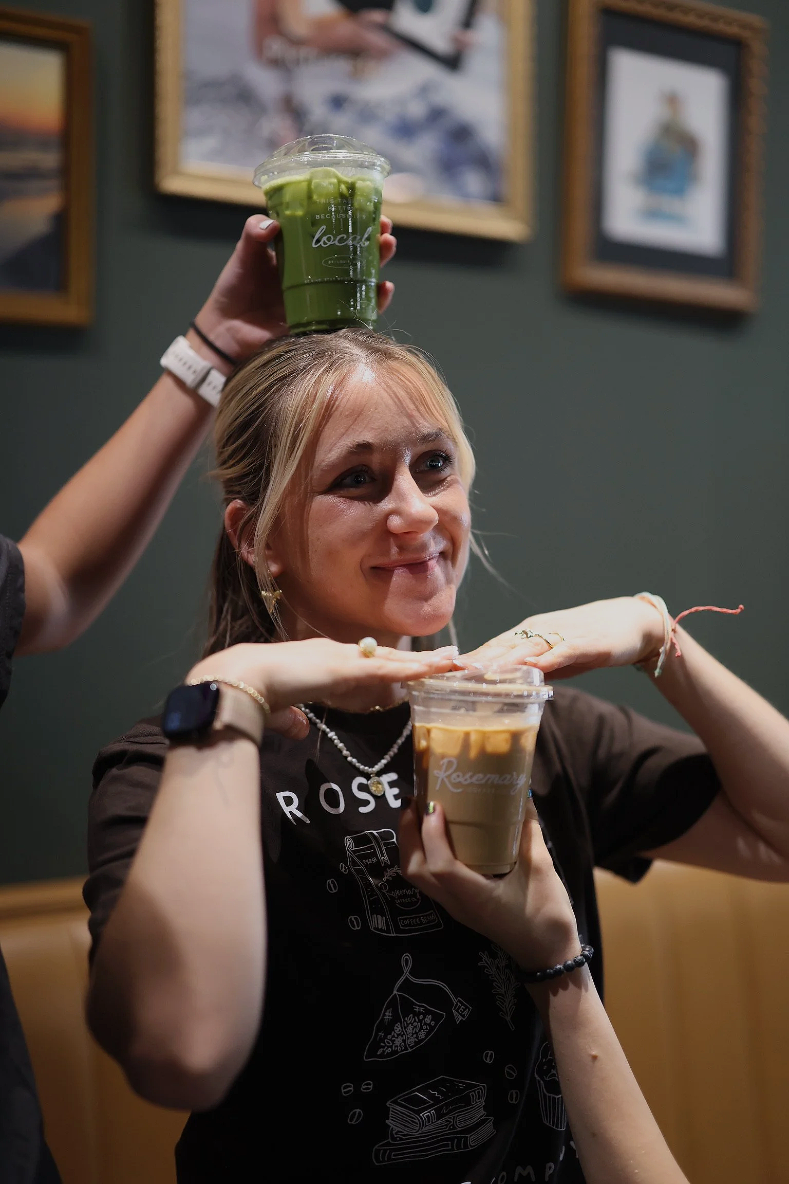 A woman with blonde hair and a black shirt is holding a plastic cup of iced coffee, and another person is holding a cup of green matcha latte above her head in a cafe with framed pictures on the wall.