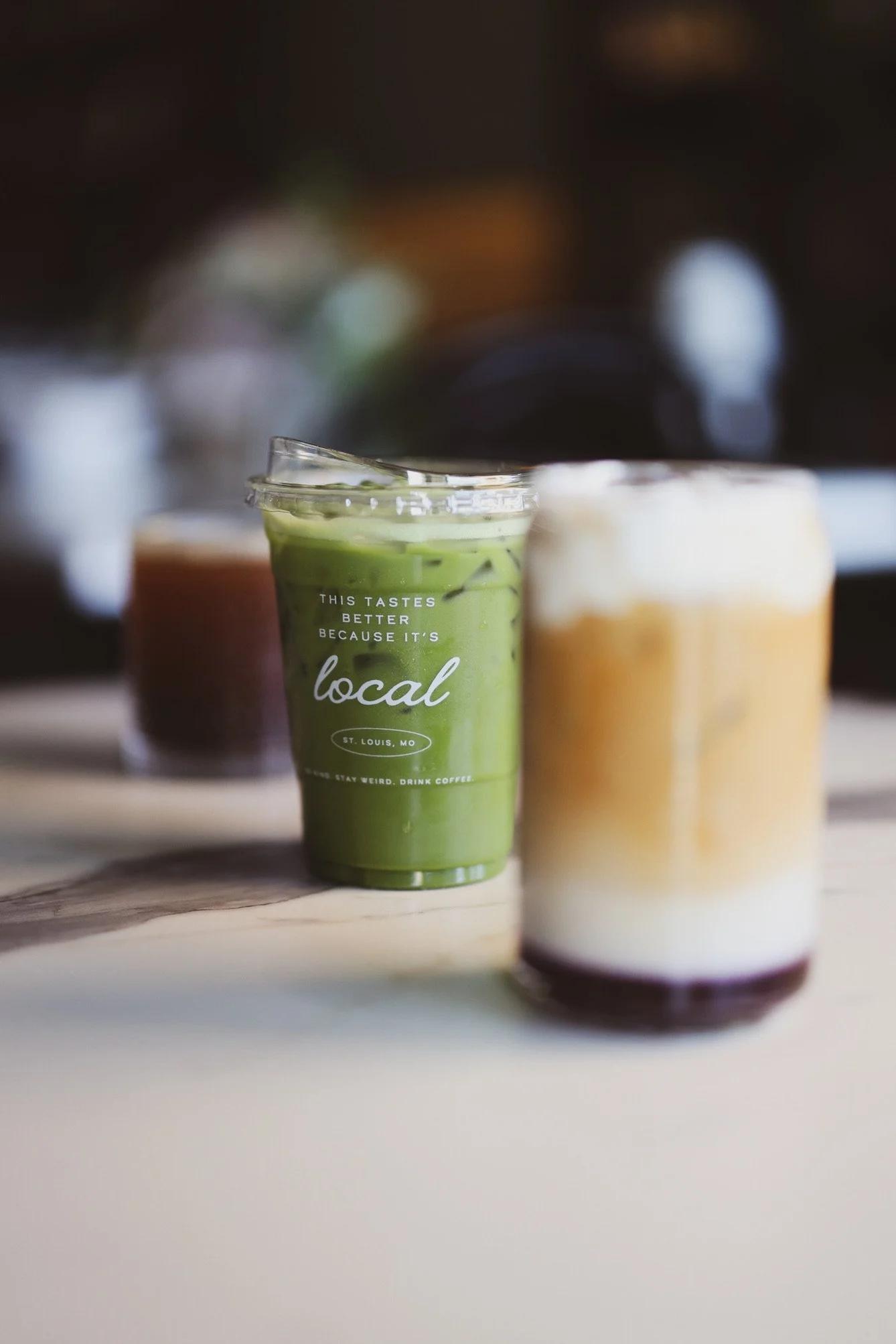 A green smoothie in a clear plastic cup with a lid, placed on a wooden board. The cup has text that says, 'This tastes better because it's local,' and 'St. Louis, MO.'