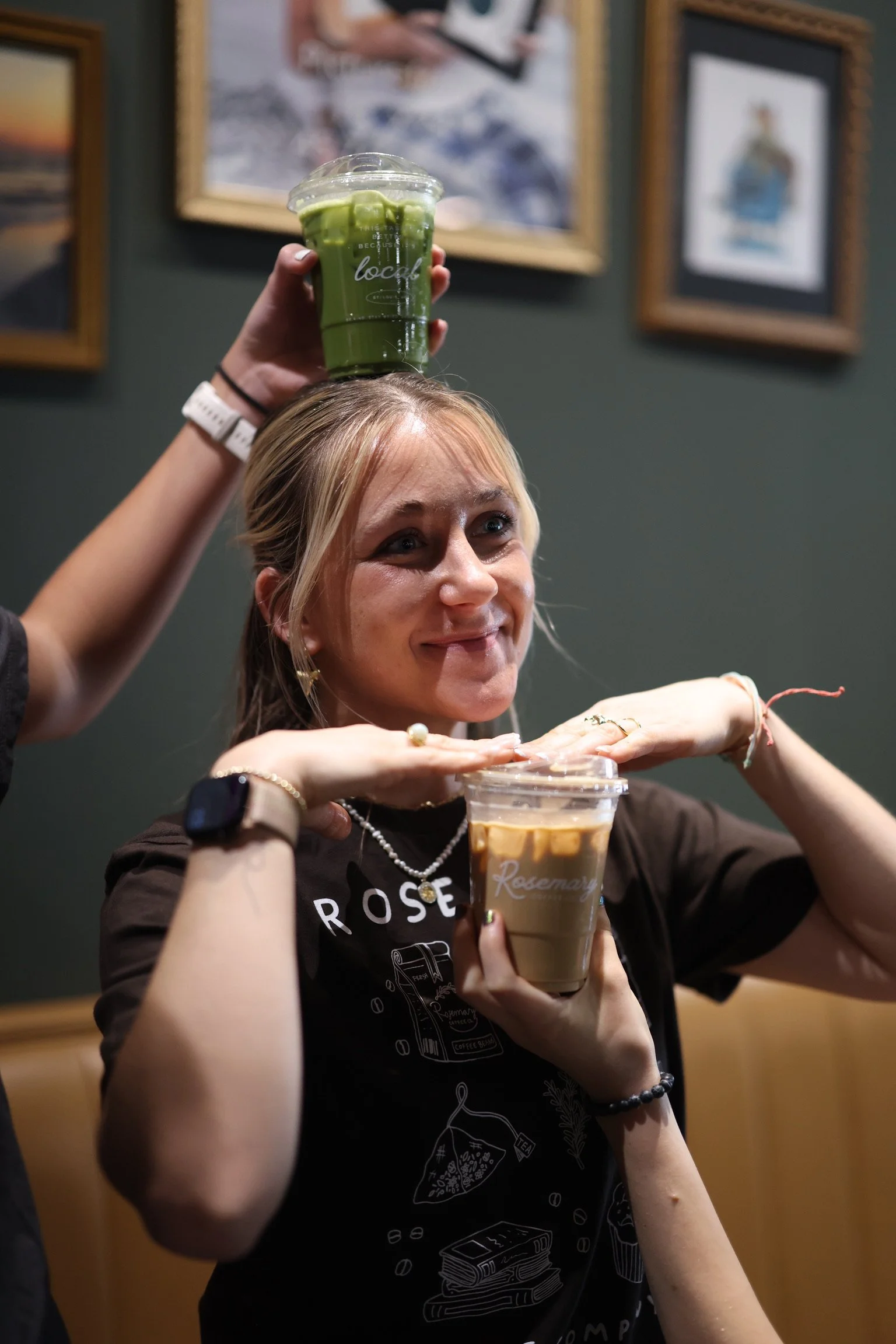 A woman with blonde hair smiling, holding a large iced coffee with a clear cup and lid, in a cozy indoor setting with framed artwork on the wall behind her.