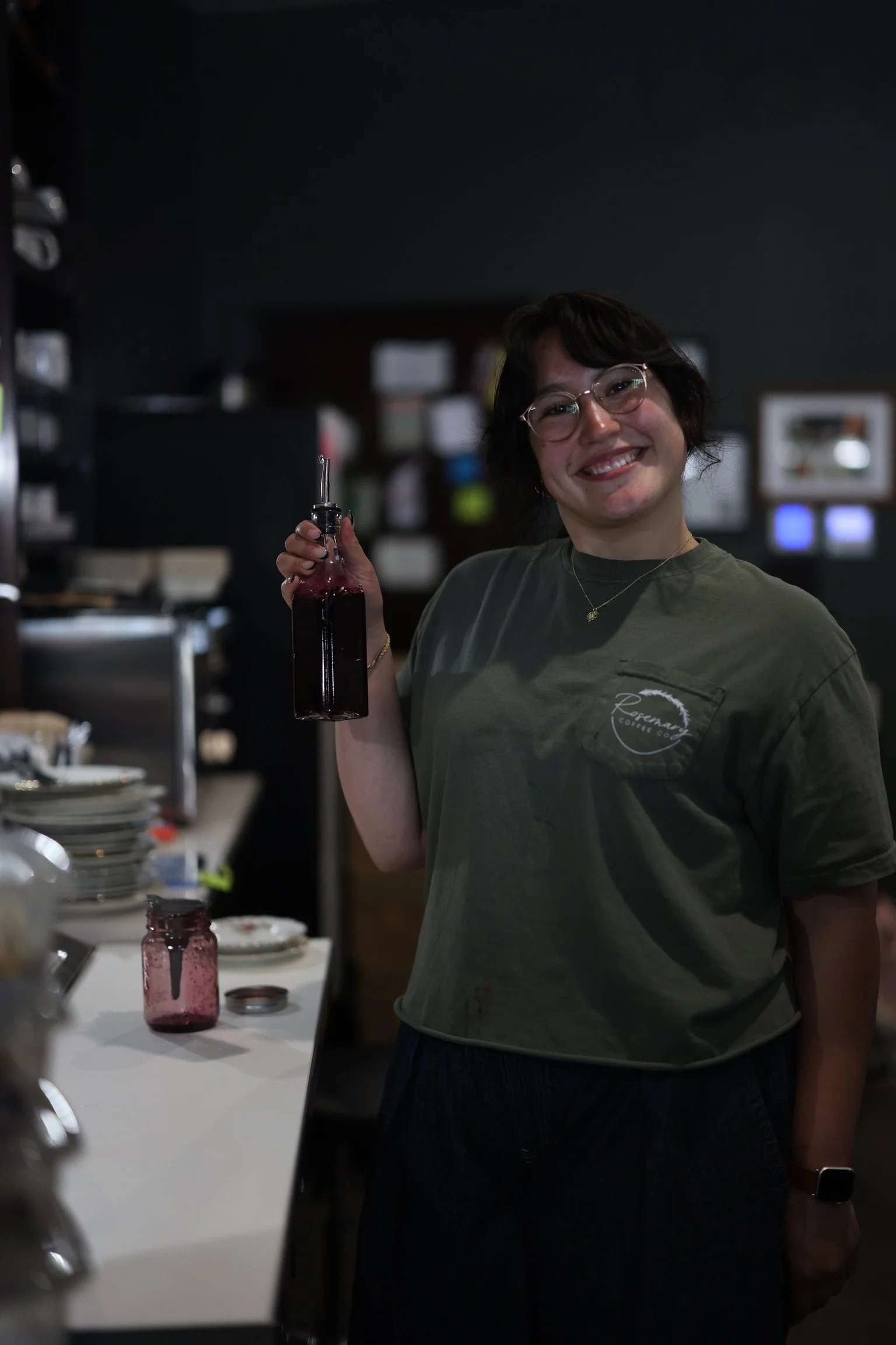 A woman with short dark hair, glasses, wearing a green T-shirt and dark pants, holding a bottle of red liquid with a syringe. She is smiling and standing in a room with plates, jars, and various kitchen items on the counter.