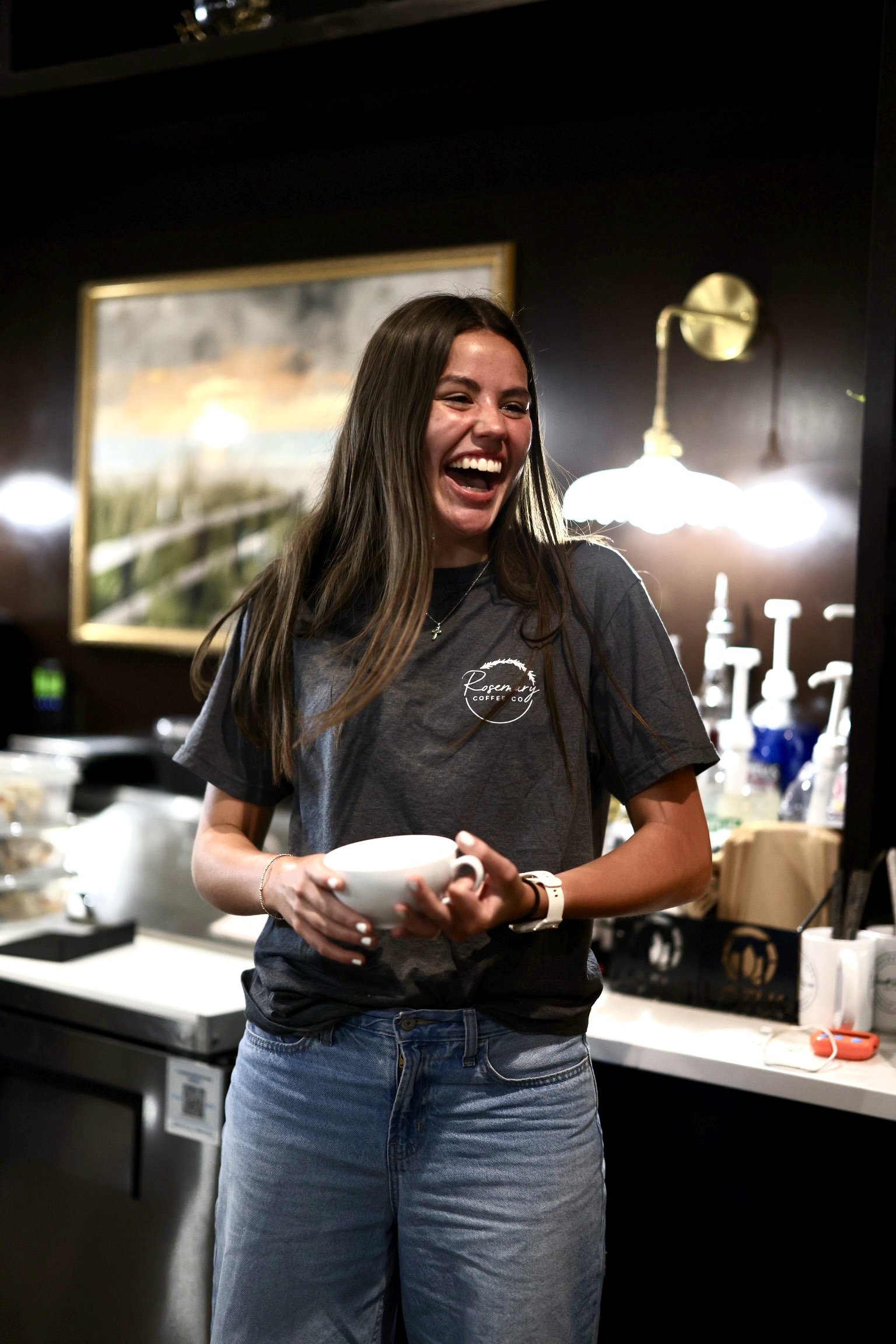 A young woman with long dark hair laughing while holding a white coffee mug in a coffee shop. She is wearing a dark gray t-shirt with white text and blue jeans. The background includes a framed picture, a wall-mounted lamp, and a counter with various items.