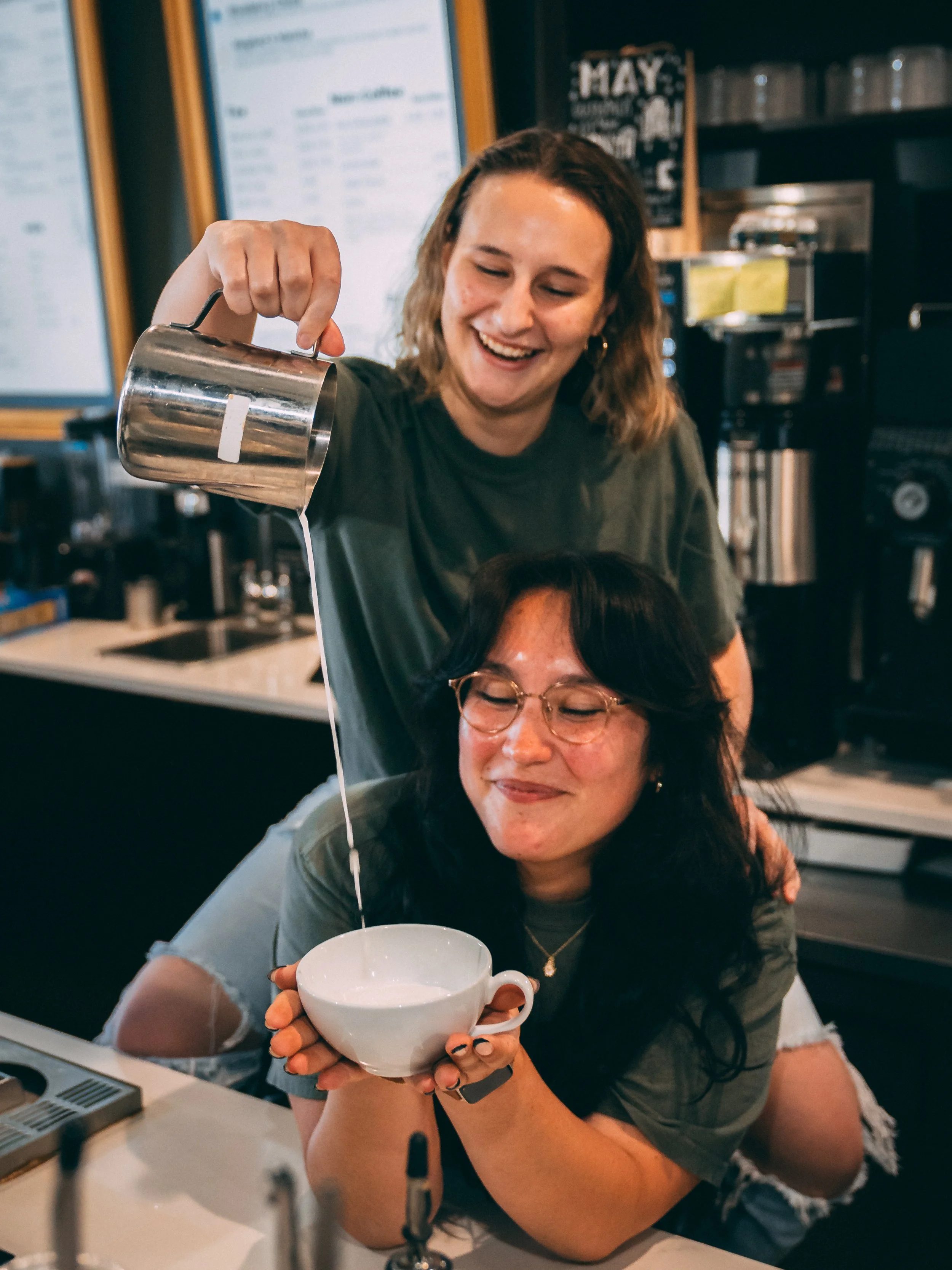 A woman in glasses holds a white mug with her eyes closed and a smile. Another woman with wavy hair, earrings, and a watch is pouring milk from a metal pitcher into the mug, smiling and looking down at her. They are in a cozy cafe setting.
