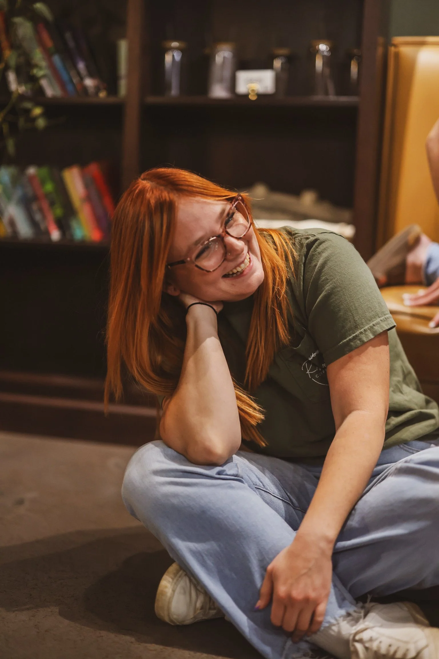 A woman with long red hair and glasses laughing, sitting on the floor with crossed legs, in a room with dark wooden shelves filled with books and jars in the background.