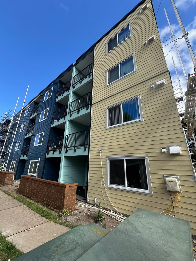 Exterior view of a multi-story residential building with blue and beige siding, multiple windows, balconies with black railings, and some construction scaffolding on the side under a blue sky.