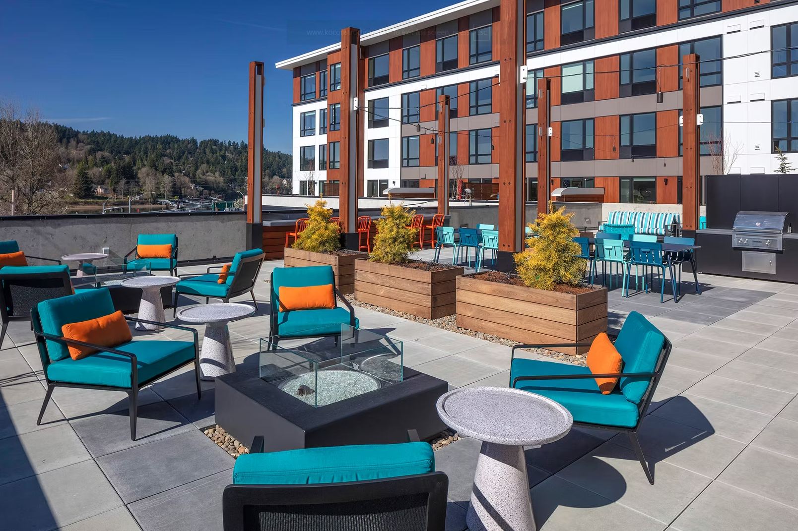 Rooftop patio with seating, planters, pergolas, a barbecue grill, and a modern apartment building in the background.