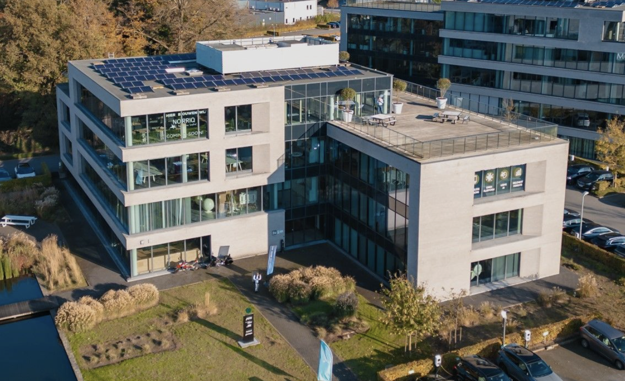 Luchtfoto van een modern kantoorgebouw met zonnepanelen op het dak, omgeven door parkeergelegenheid en tuinruimte, in een stedelijk gebied met andere kantoorpanden.