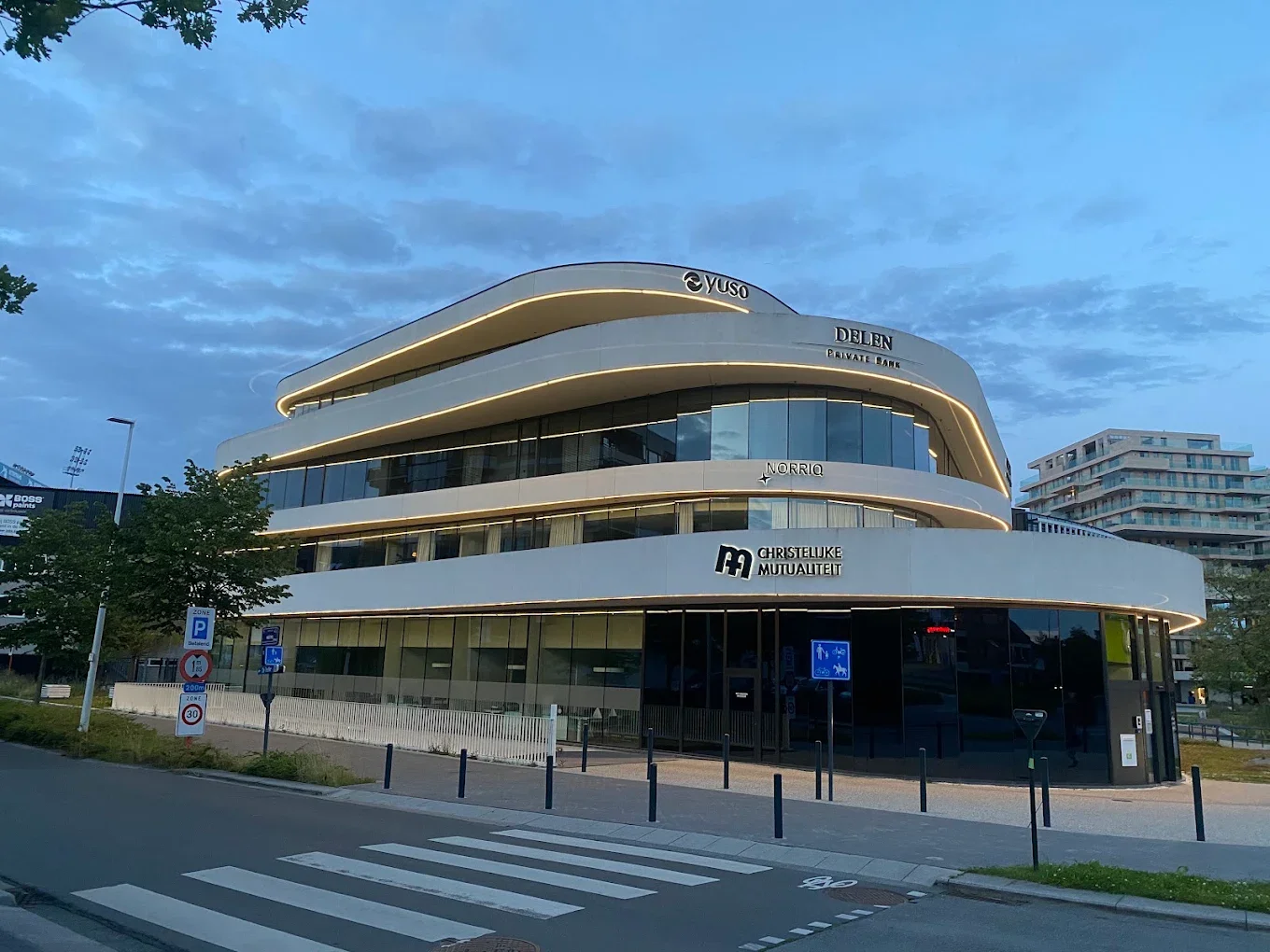 Modern wit kantoorgebouw met glanzende ramen en ronde architectuur, met logos en hoge verlichting langs de randen, omringd door een weg met voetgangerspaden en verkeersborden.
