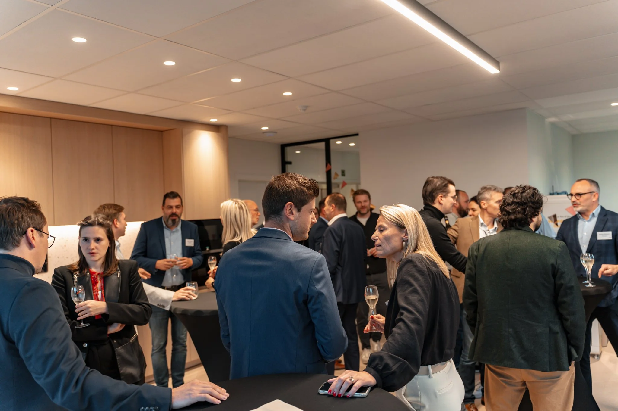 Een groep mensen op een netwerkevenement of zakelijke bijeenkomst, pratend en drinkend in een moderne binnenruimte.