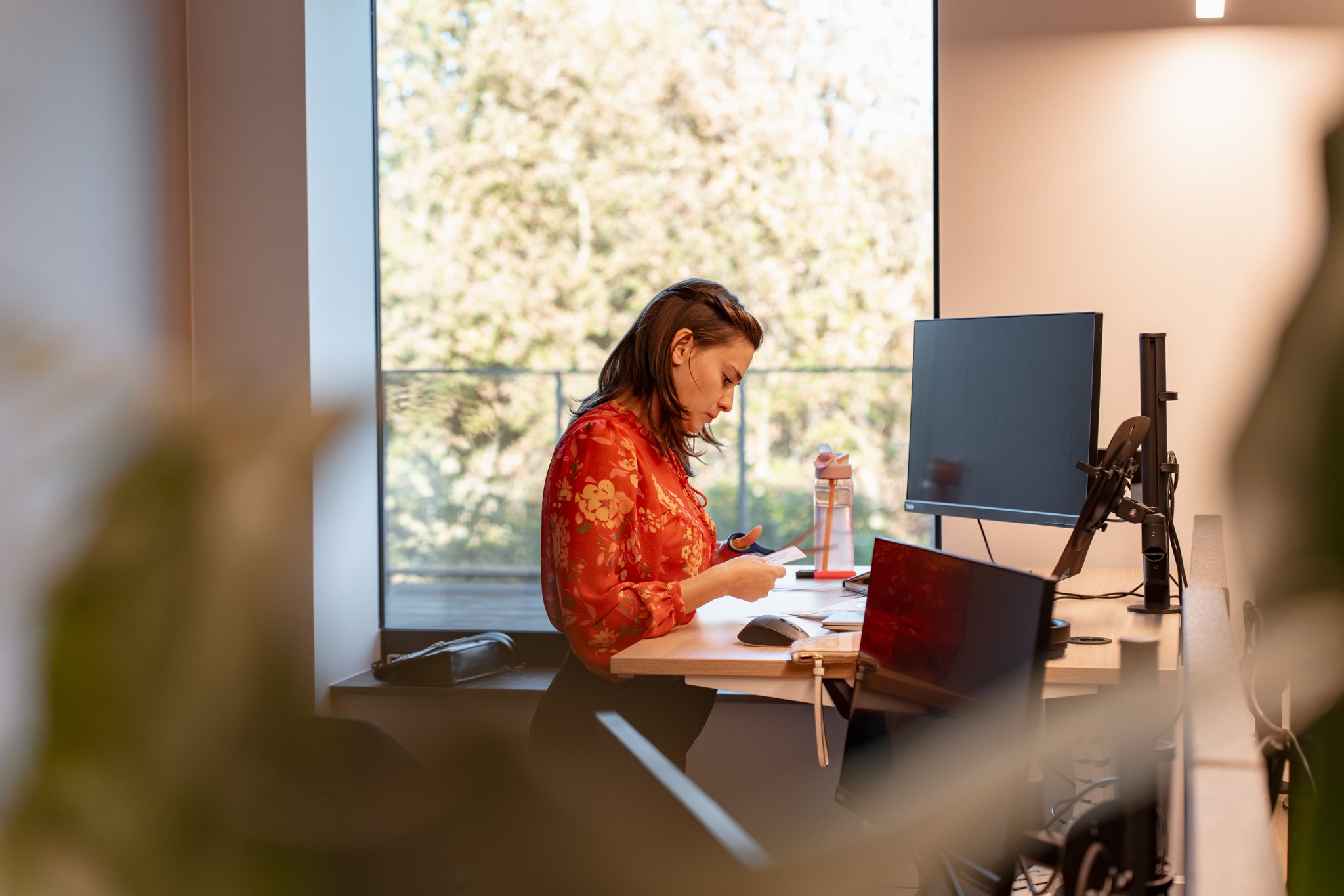 Vrouw zit aan een kantoorwerkplek, kijkt naar haar telefoon, met een computerscherm en andere kantoorbenodigdheden op haar bureau, bij daglicht door een grote raam met uitzicht op bomen.