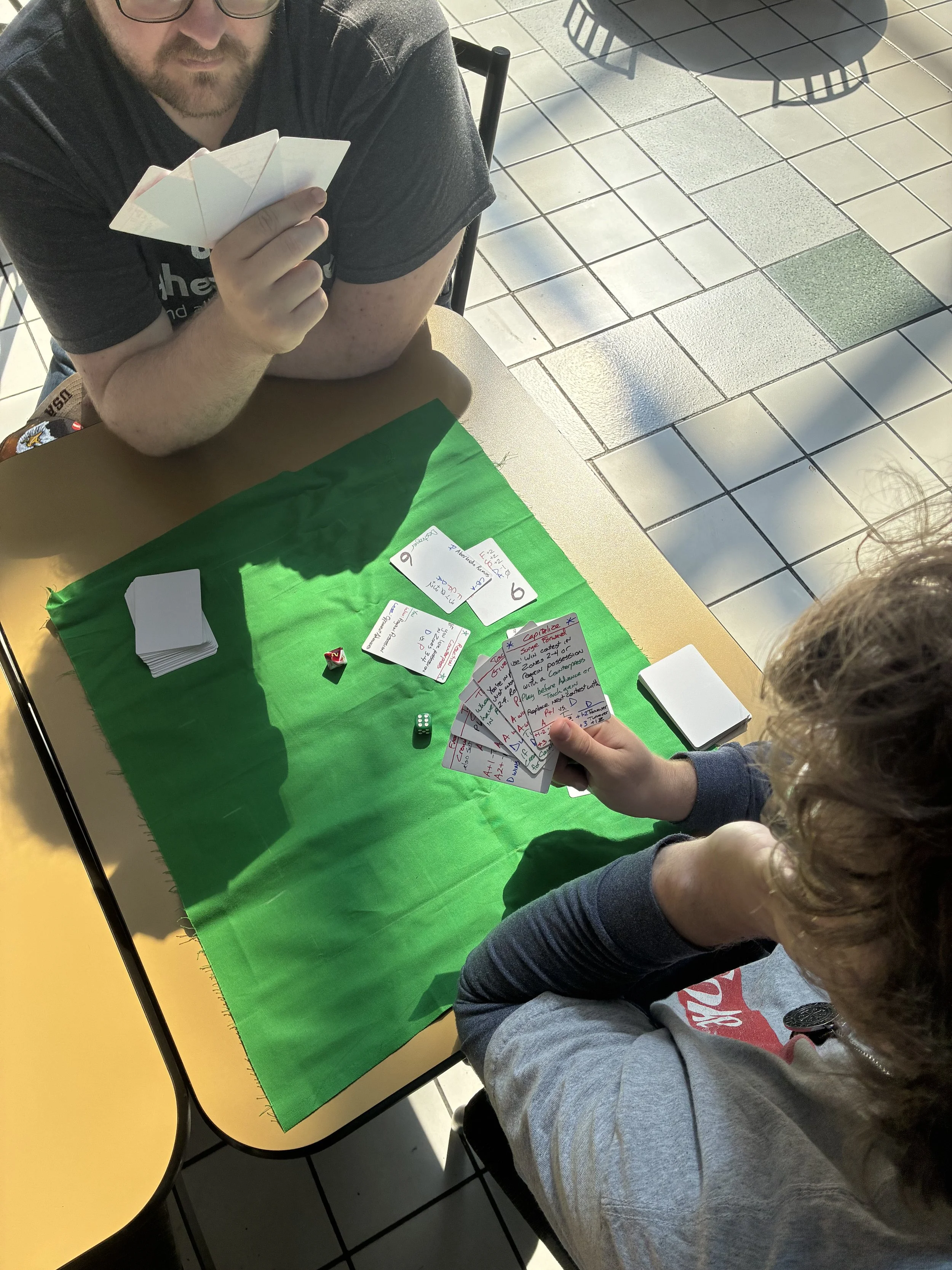 A person sitting at a table with a green cloth, playing a card game with another person whose hand is visible holding several cards. There are dice and notes on the table, and the setting appears to be a tiled indoor or outdoor space with chairs and shadows.