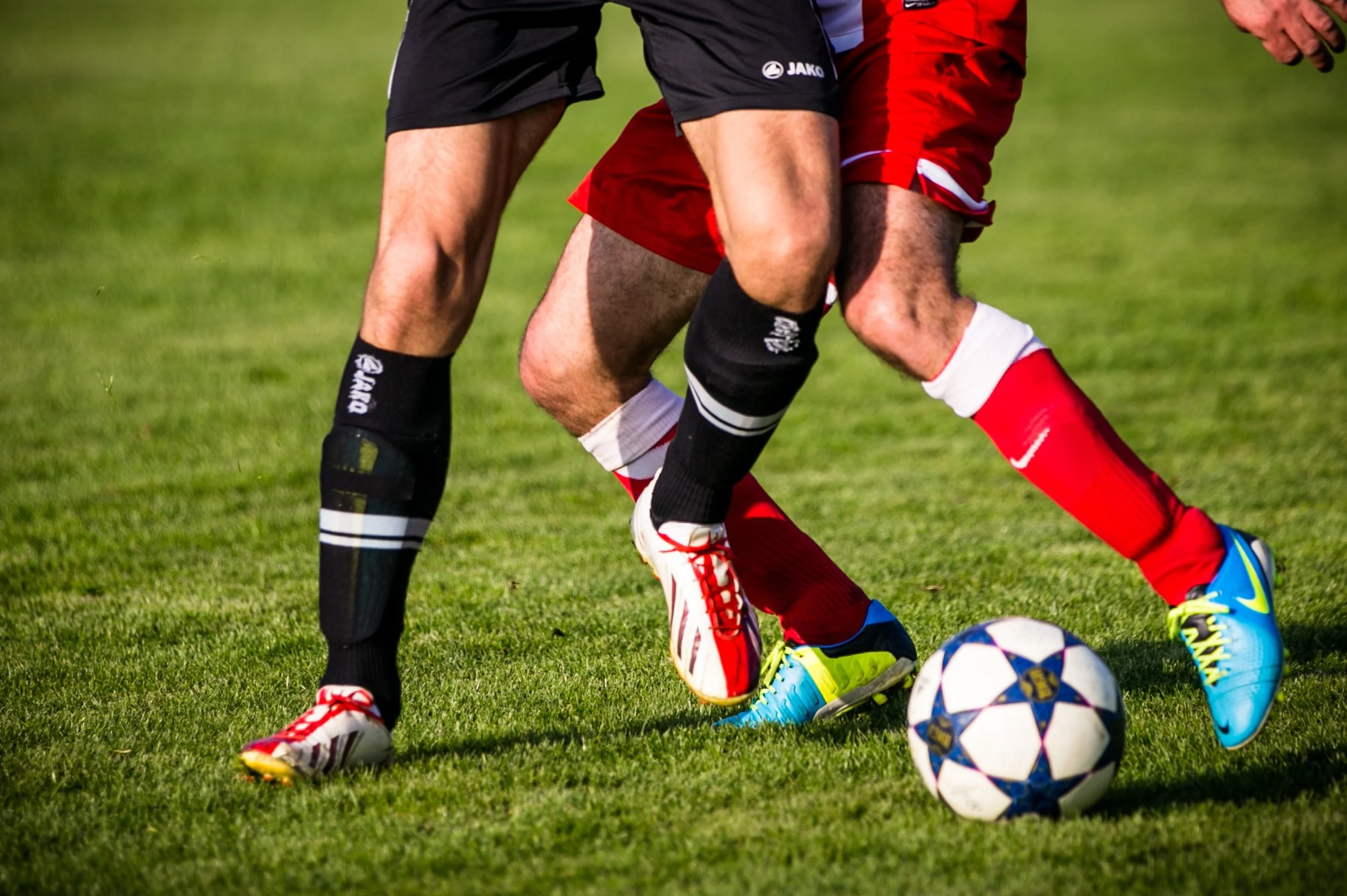 Two soccer players competing for the ball on a grassy field, one in black and the other in red shorts, with a soccer ball and colorful cleats visible.