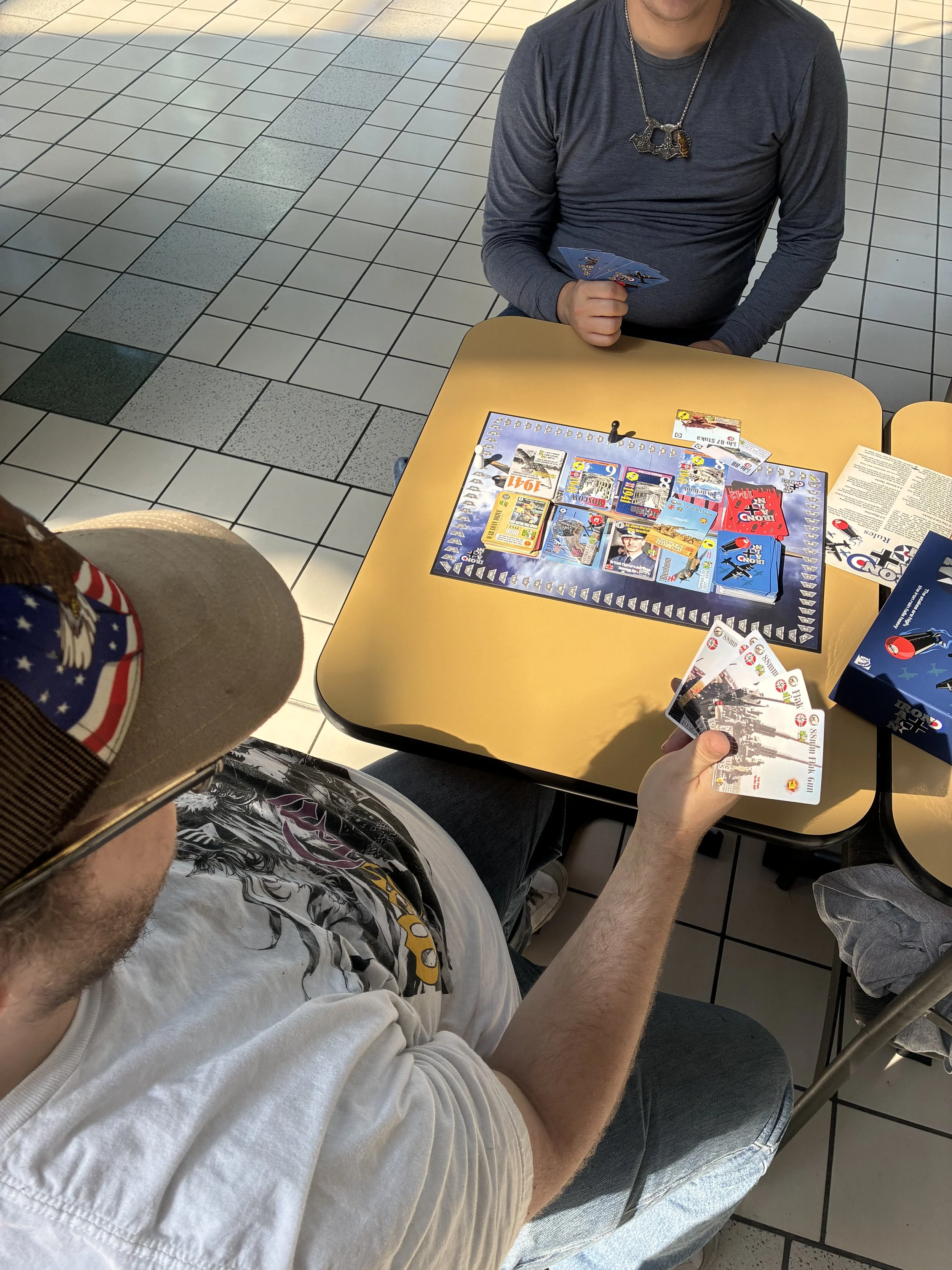 Two people sitting at a table playing a card game. One person is holding cards, and the other is holding fan of cards. The table has a board game with various cards and game pieces on it, and there are pamphlets and boxes on the table. The floor is tiled with gray and white tiles.