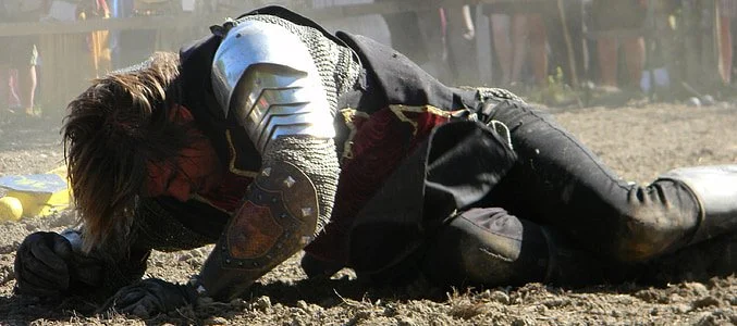 Person dressed as a medieval knight lying on the ground during a reenactment or event.