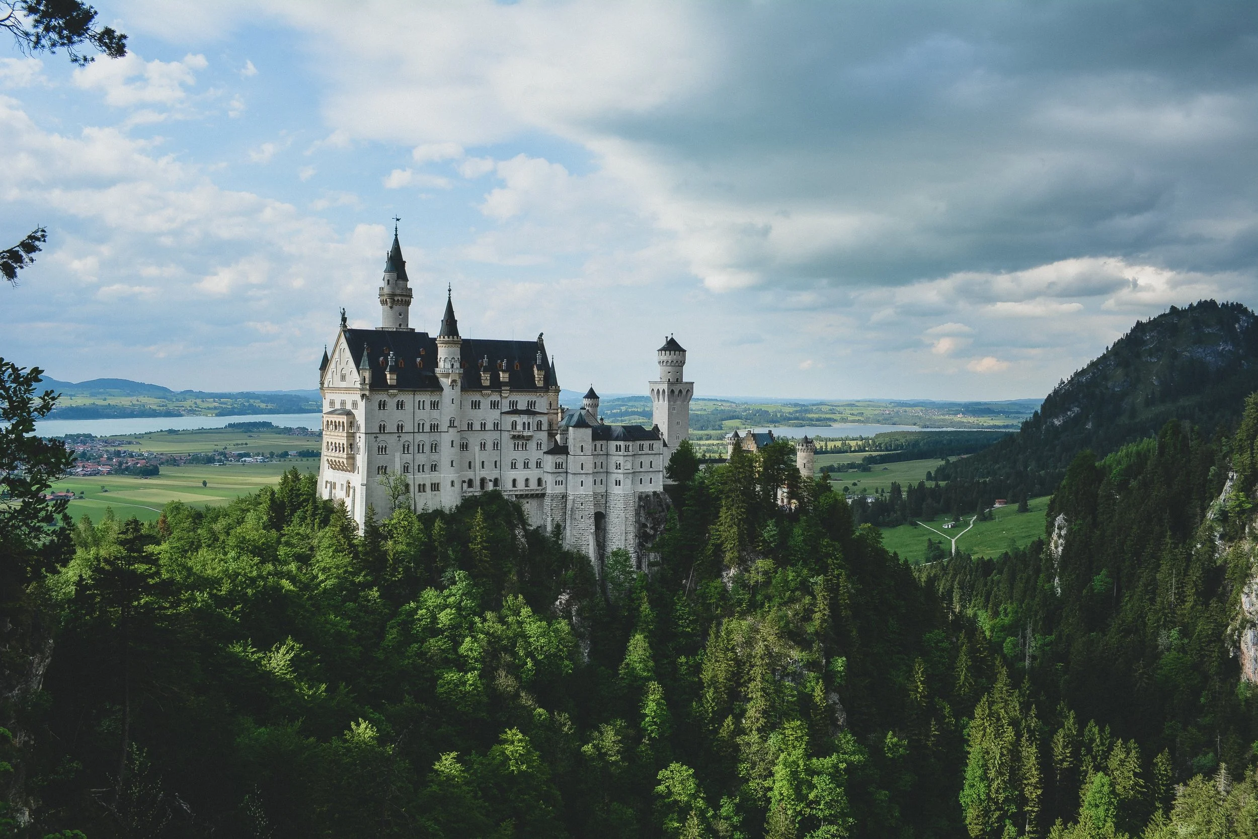A scenic view of a castle with tall towers and spires on a hill surrounded by green trees, with a body of water and a distant landscape in the background under a partly cloudy sky.