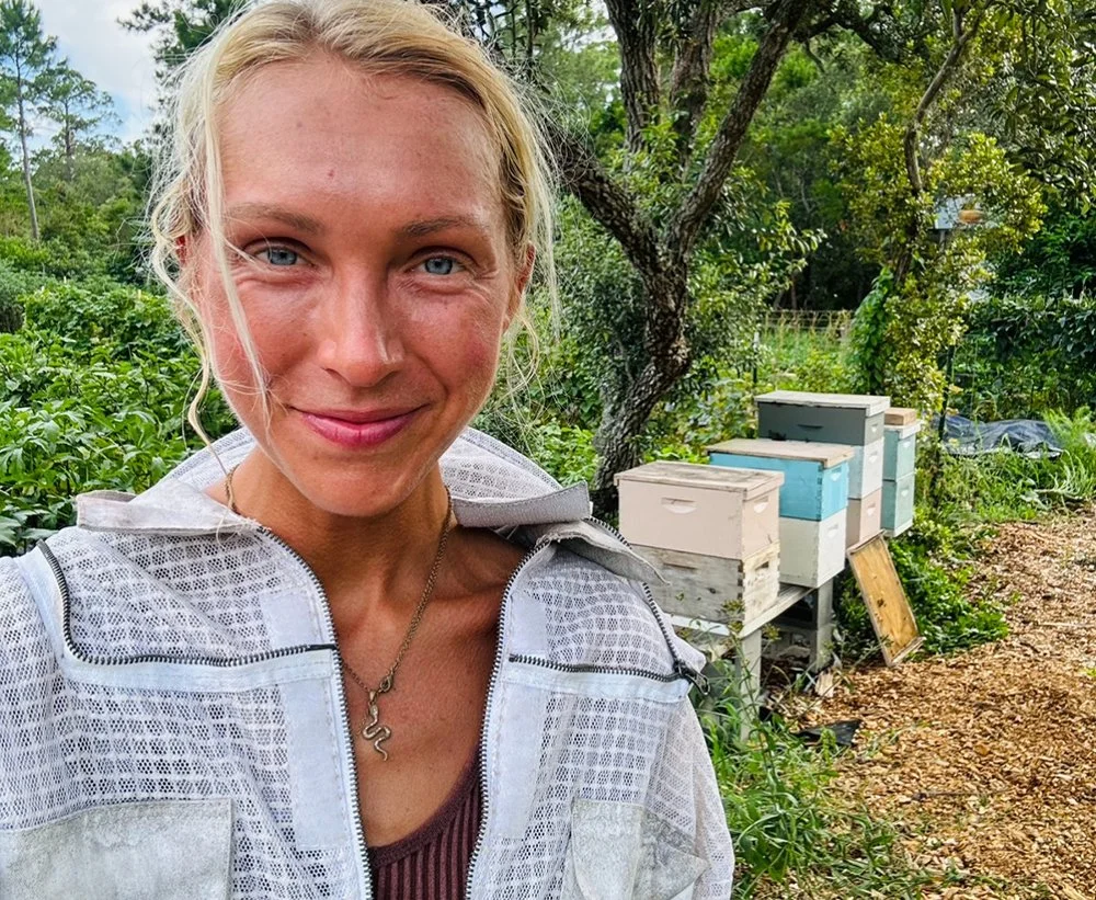 A smiling woman with blonde hair and blue eyes stands outdoors in front of a lush garden with beehives on the right side.